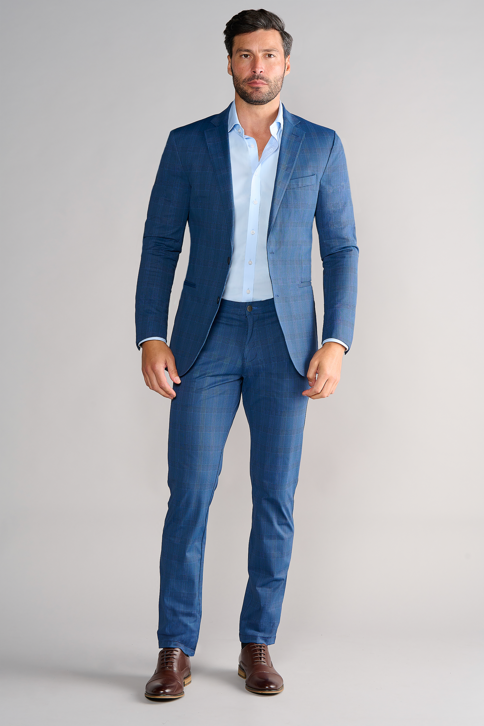 A man with short brown hair and a trimmed beard wears the Twillory Performance Blazer in Blue Prince of Wales over a light blue dress shirt, paired with brown dress shoes. He stands facing forward against a plain light gray background.