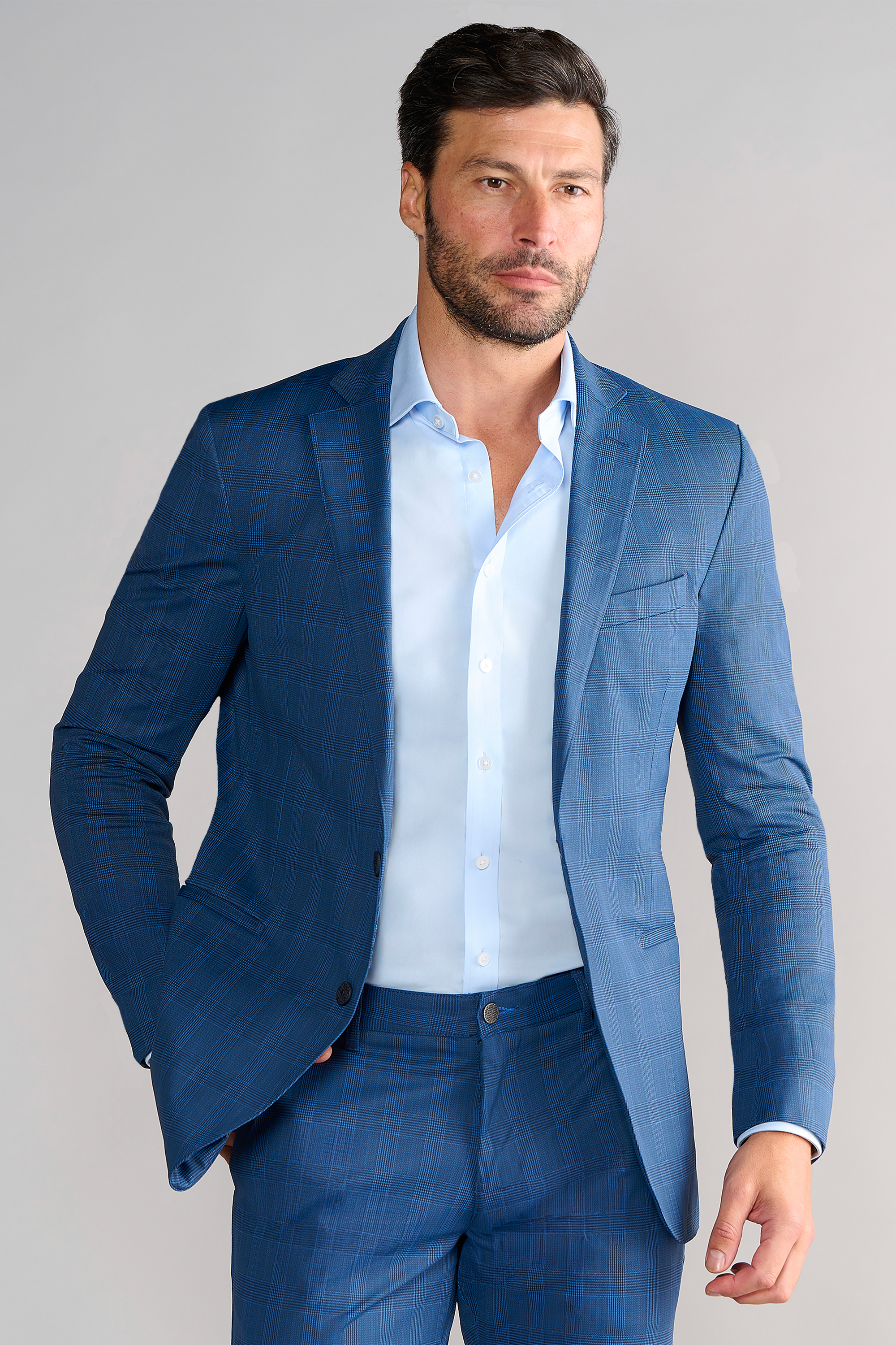 A man with short dark hair and a beard wears the Twillory Performance Blazer in Blue Prince of Wales over a light blue shirt, his right hand in his pocket, looking to the side against a plain light background.