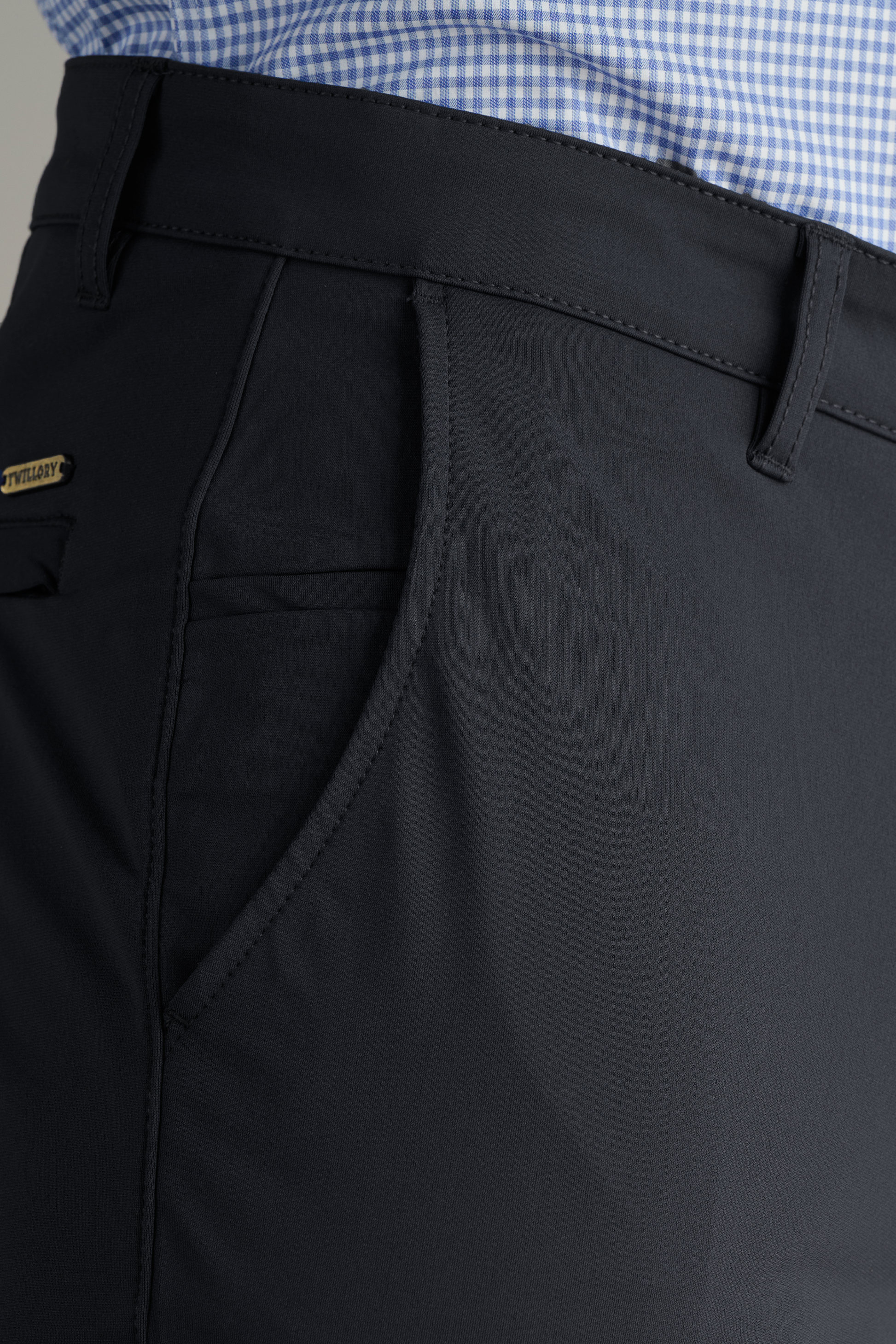 Close-up of a person wearing Twillory Performance Pants // Charcoal with a visible side pocket. The tucked-in blue and white checkered shirt highlights the pants' stitching and textured performance fabric.