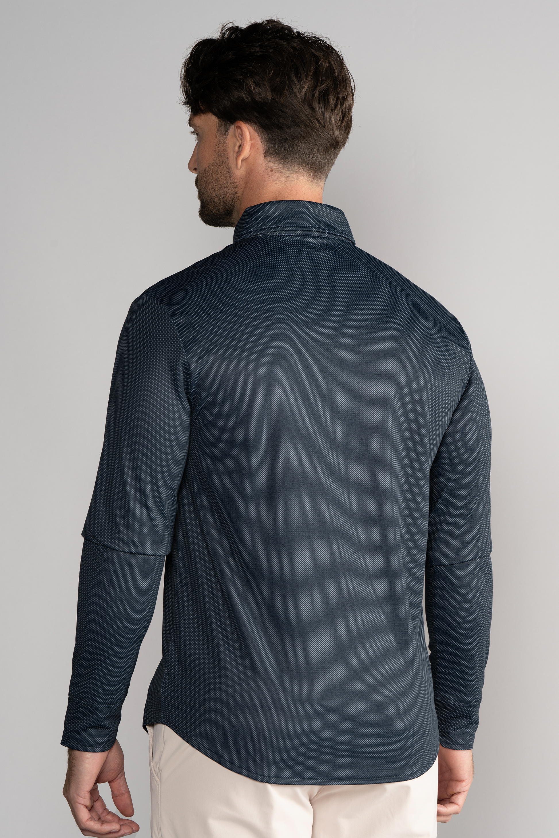 A man faces away from the camera, wearing Twillory’s Long Sleeve Button Down Polo Dot in Charcoal and light beige pants. The shirt features a subtle dot texture, a collar, moisture-wicking comfort, and is set against a plain light background.