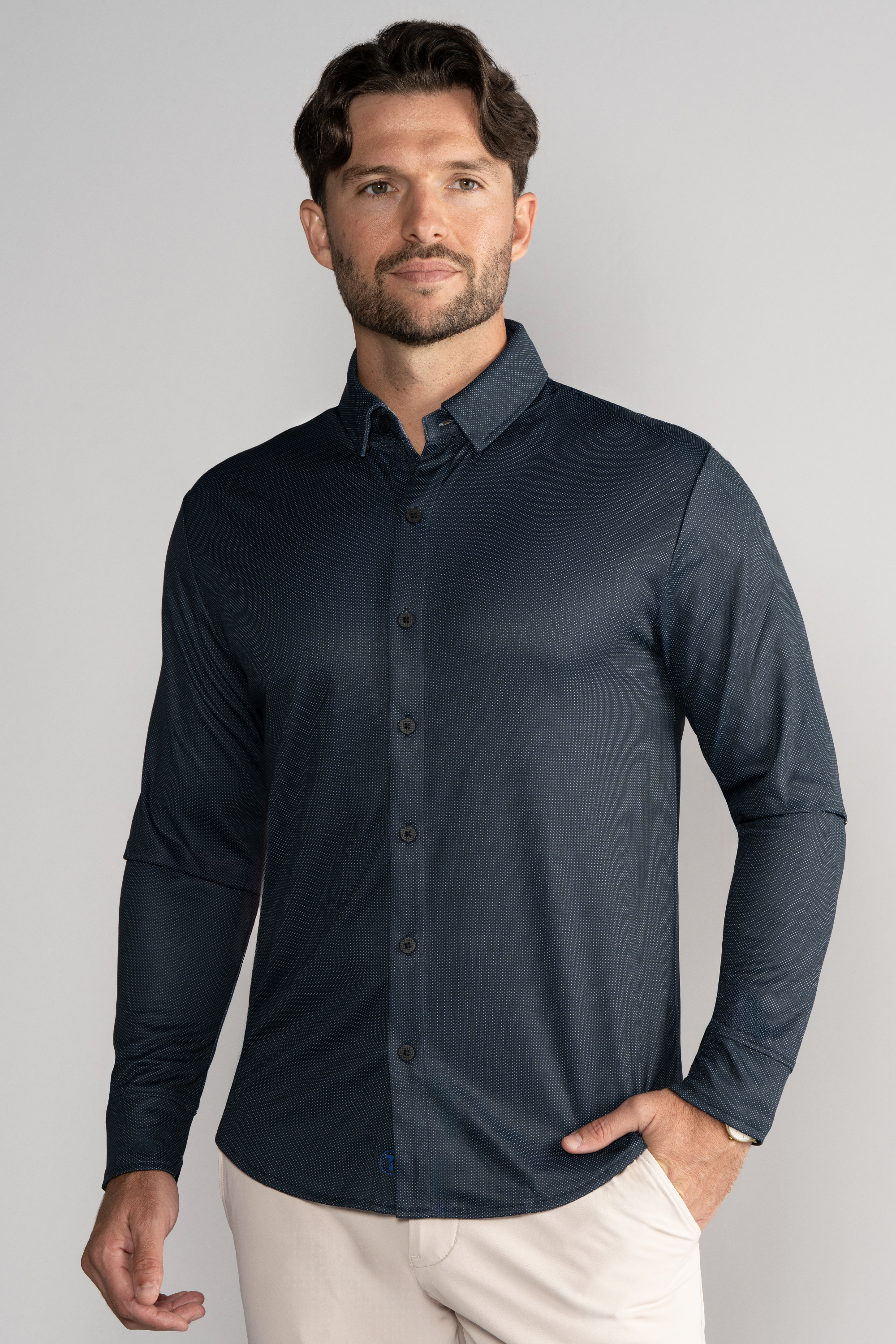 A man with short brown hair and a beard wears the Twillory Long Sleeve Button Down Polo Dot in Charcoal, paired with light beige pants. He stands facing forward with one hand in his pocket against a plain light gray background, looking neutral.