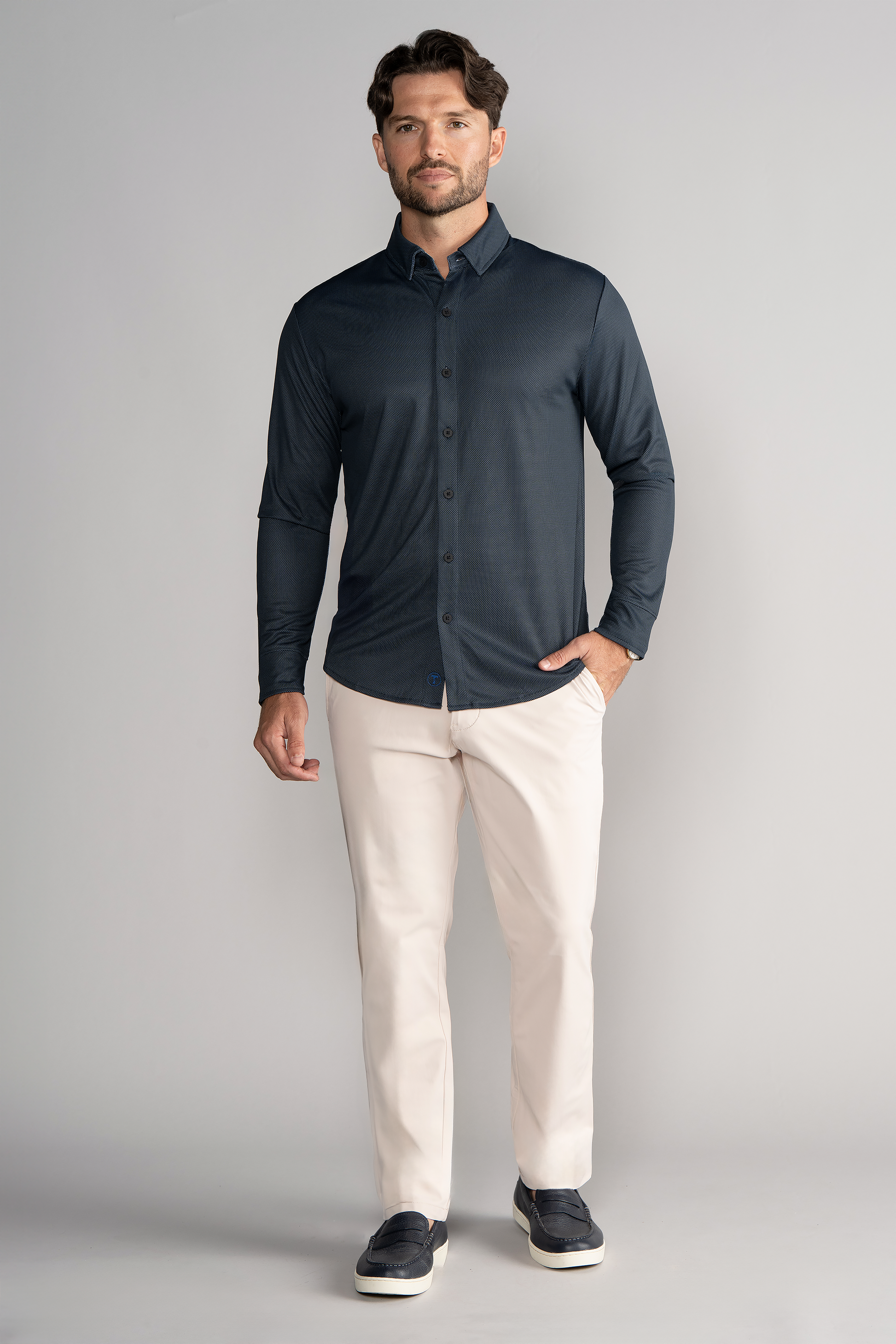 A man with short brown hair, a beard, and a neutral expression stands against a plain light gray background wearing the Twillory Long Sleeve Button Down Polo Dot in Charcoal. One hand is relaxed by his side.