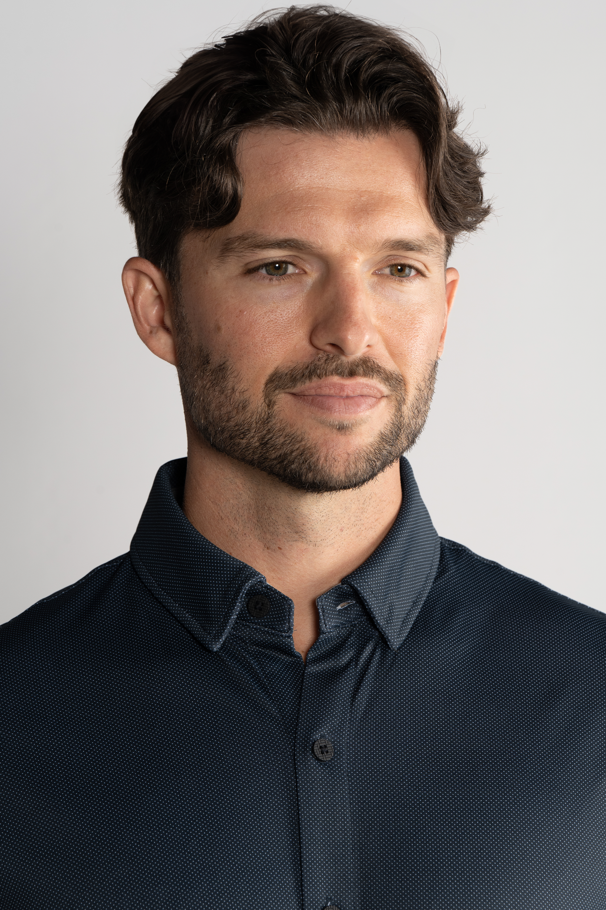 A man with short, wavy brown hair and a trimmed beard wears the Twillory Long Sleeve Button Down Polo Dot in charcoal. He faces slightly right, looking ahead with a gentle, neutral expression against a plain light background.