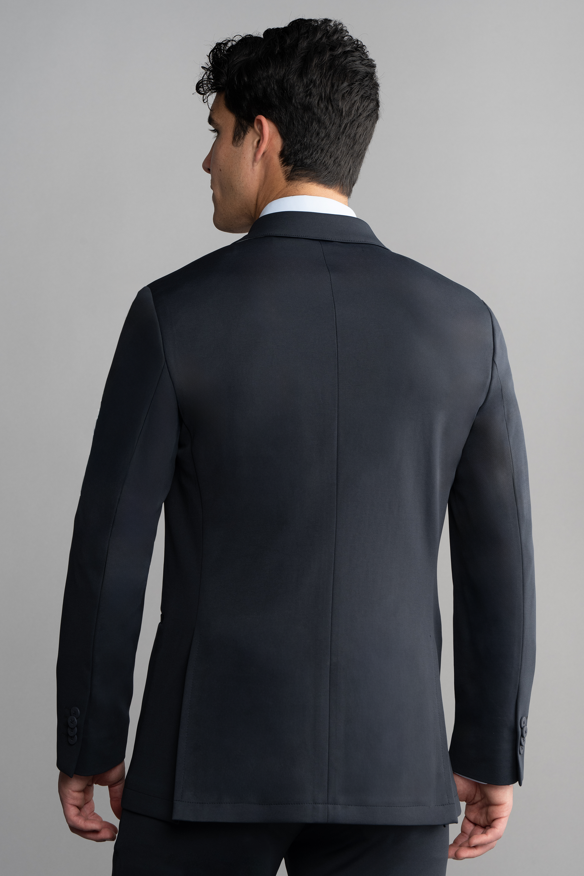 A man with short, dark hair wears the Twillory Circular Knit Blazer in black over a white dress shirt. He stands with his back to the camera against a plain light gray background, head turned slightly to the left.