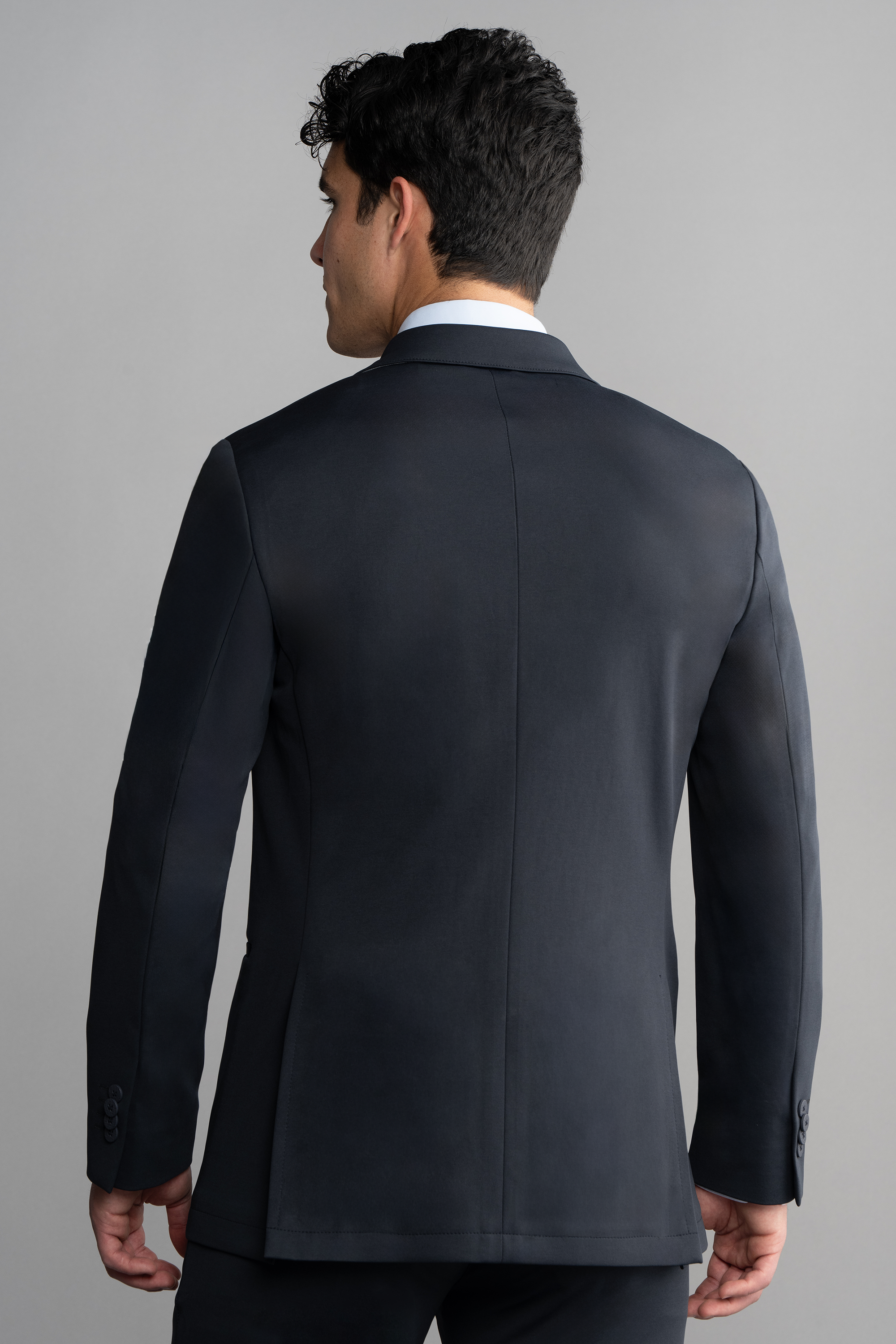 A man with short, dark hair wears the Twillory Circular Knit Blazer in black over a white dress shirt. He stands with his back to the camera against a plain light gray background, head turned slightly to the left.