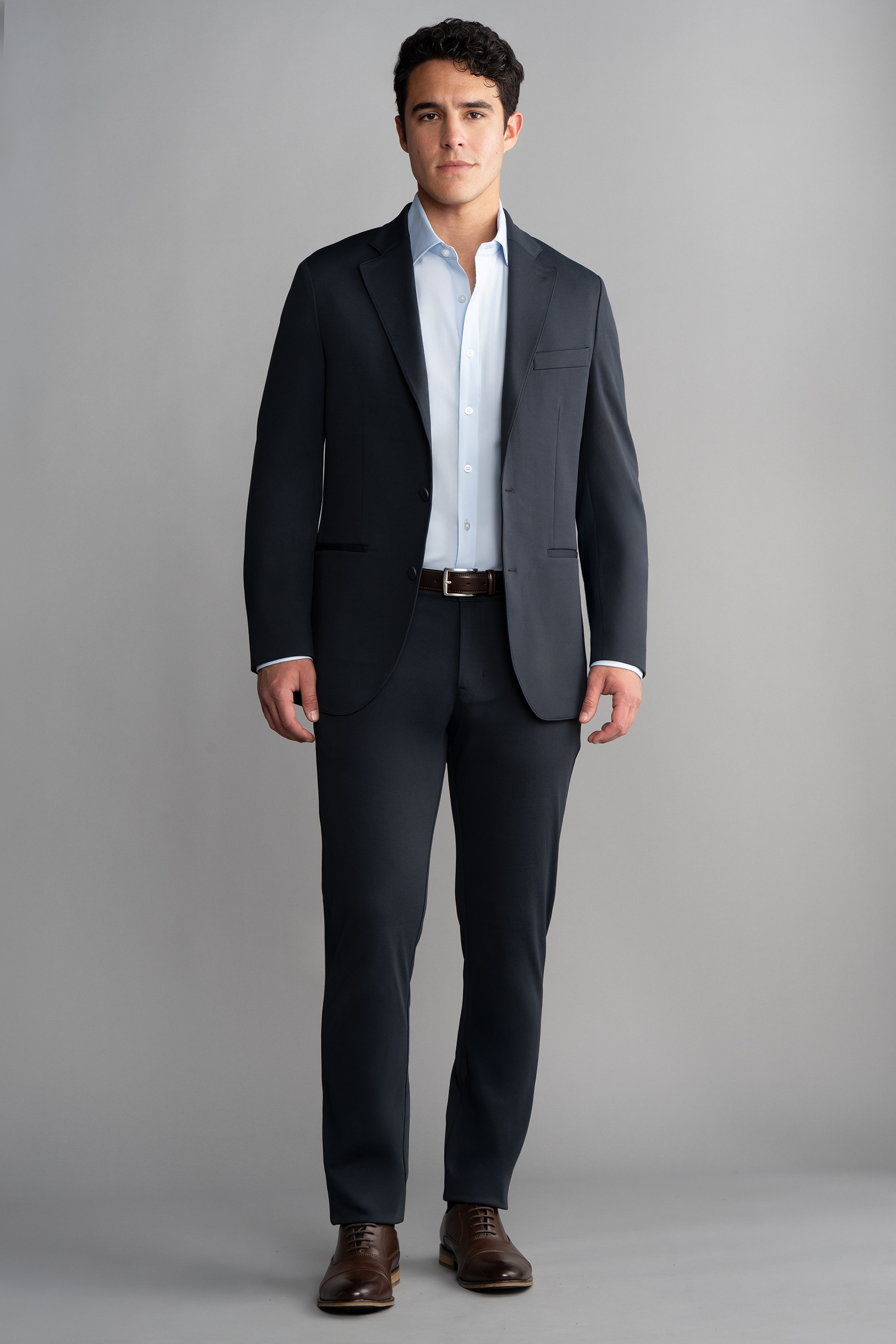 A man with short dark hair stands facing forward against a plain light gray background, wearing Twillory Circular Knit Pants // Black along with a fitted dark suit, white dress shirt, dark belt, and brown dress shoes. His hands are by his sides.