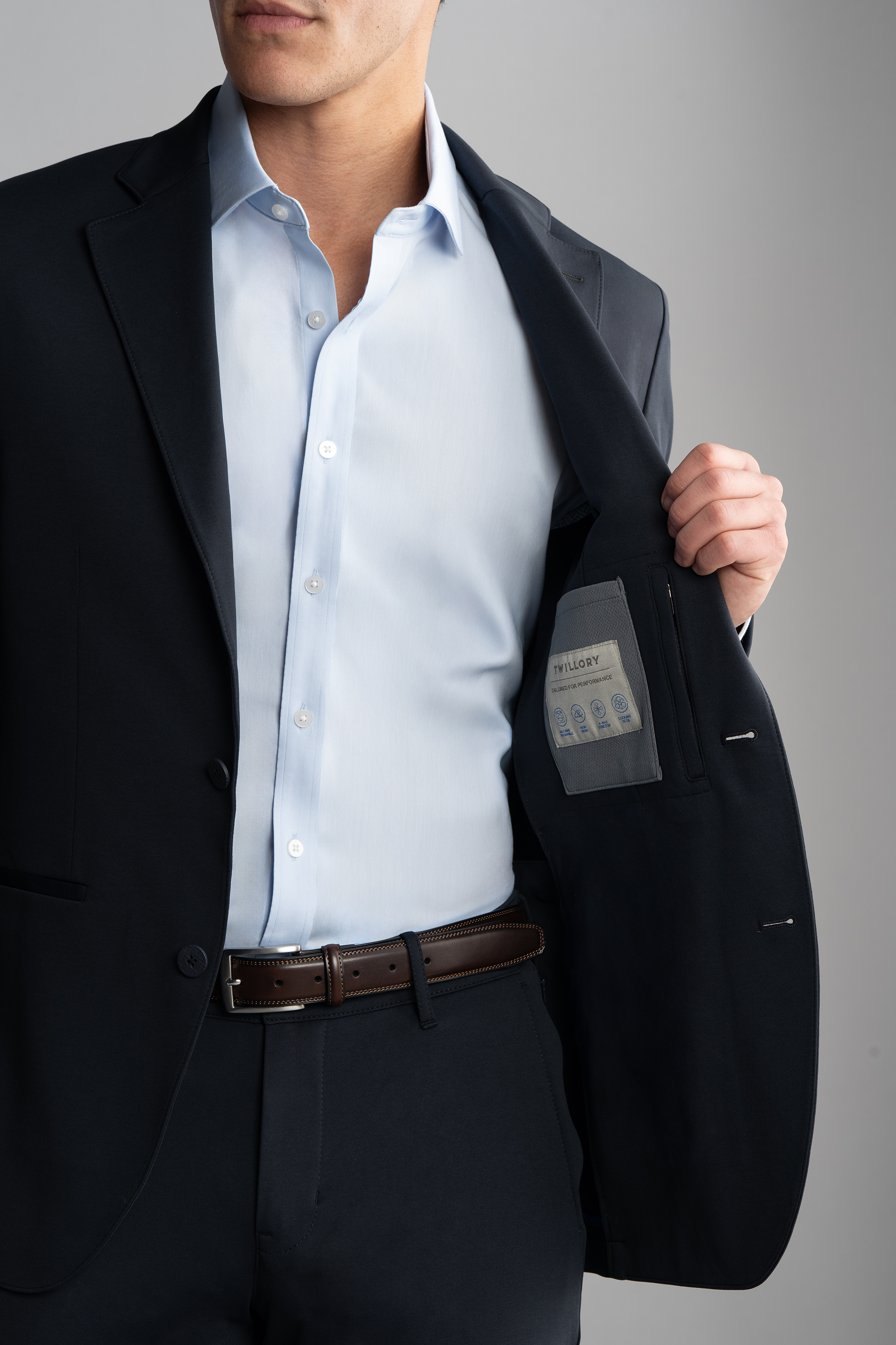 A man wearing the Twillory Circular Knit Blazer in black and a light blue shirt opens his blazer to reveal the inner pocket and label, standing against a plain gray background with a brown leather belt.