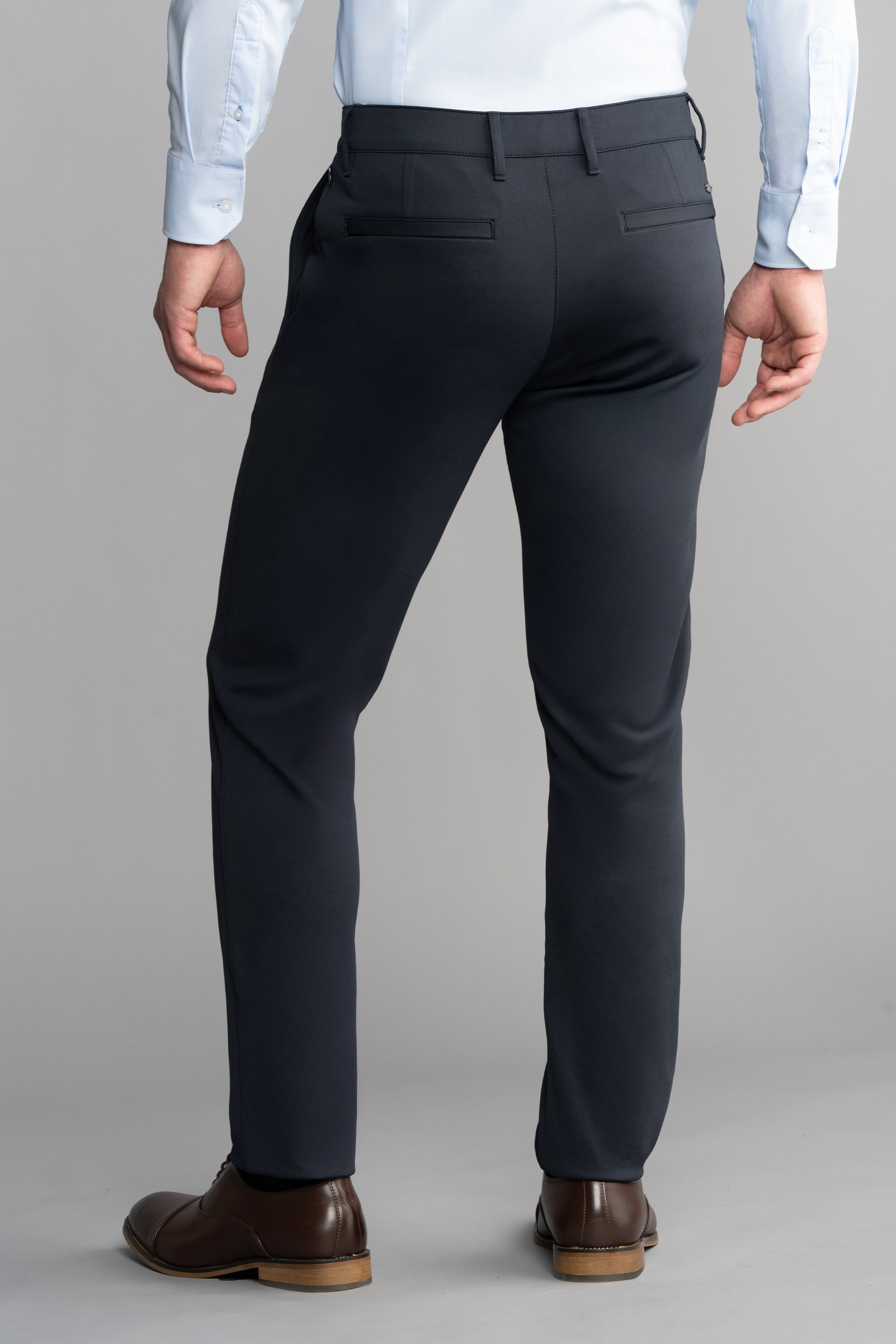 A person wearing Twillory Circular Knit Pants // Navy with 4-way stretch, a light blue dress shirt, and brown shoes stands facing away, highlighting the wrinkle-resistant welt pockets and crisp fabric against a plain gray background.