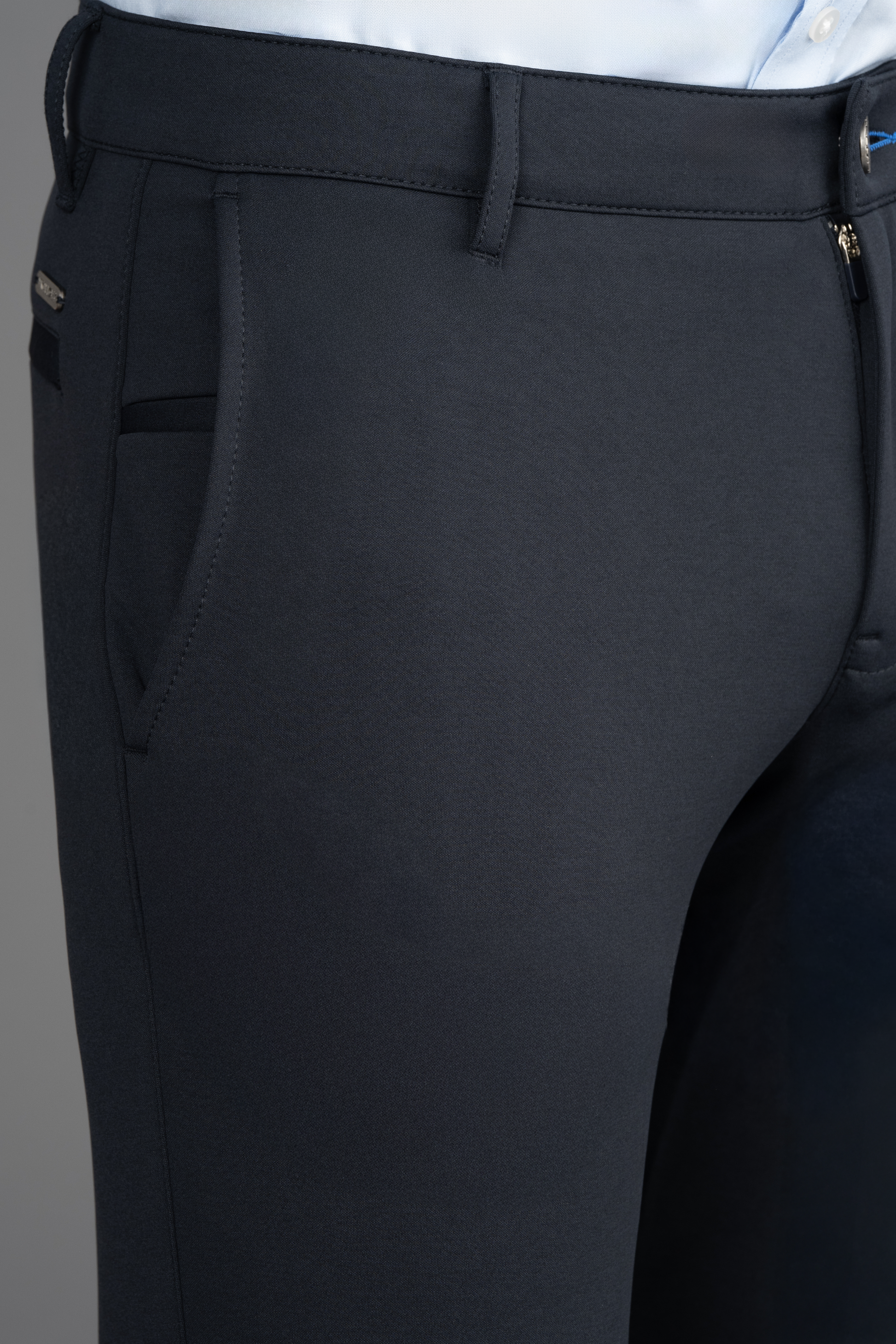 Close-up of Twillory's Circular Knit Pants in Black featuring a visible side pocket, belt loop, and zipper. A light blue shirt is tucked into the waistband. The plain gray background highlights the fabric's texture and details.
