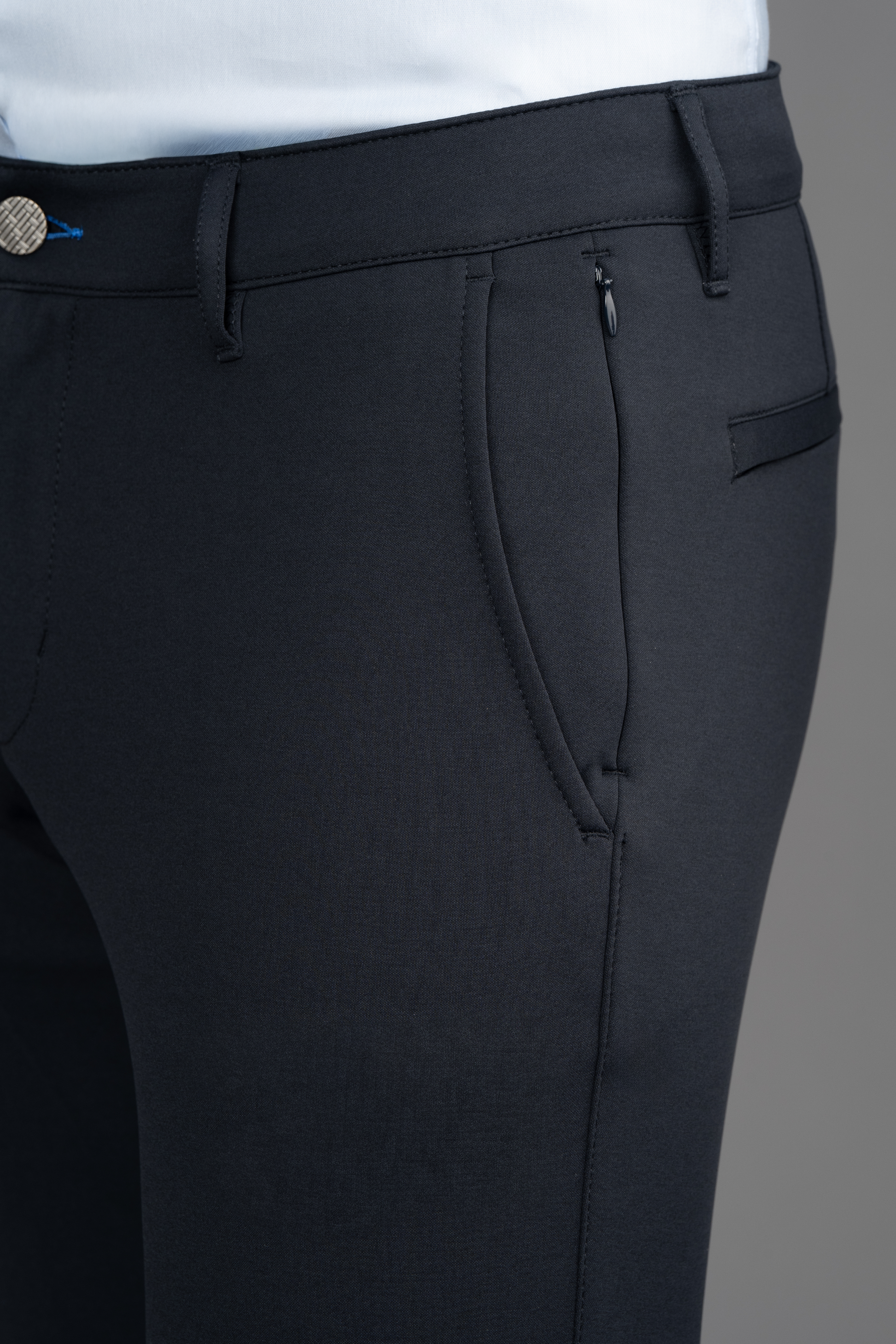 Close-up of a person wearing Twillory Circular Knit Pants // Black and a white shirt. The pants offer 4-way stretch, zippered front pocket, belt loops, side seam, and a subtle waistband button. Background is neutral gray.