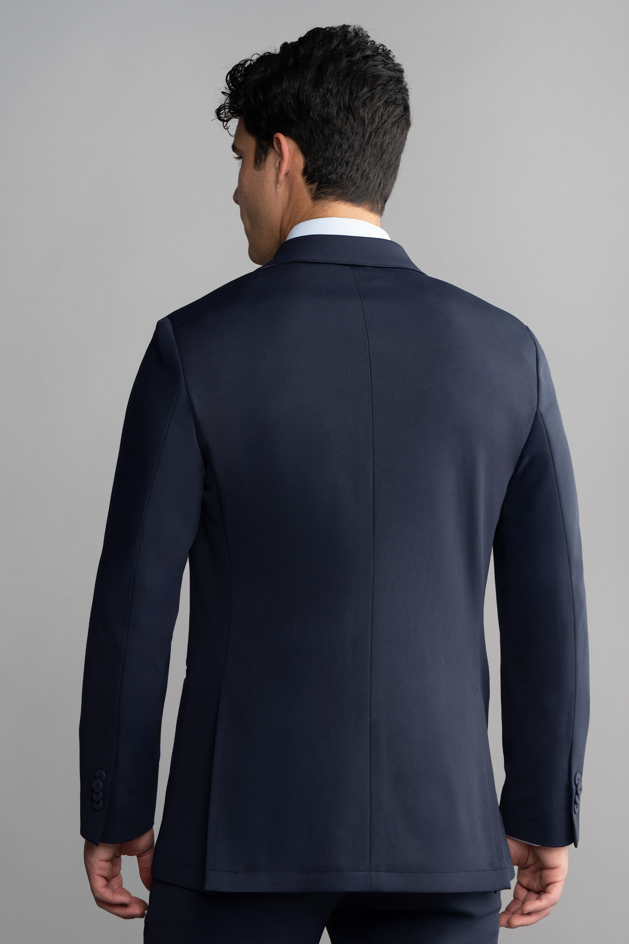 A man with short, dark, curly hair stands facing away from the camera, wearing Twillory's Circular Knit Blazer in Navy with matching trousers and a white dress shirt. The background is plain and light gray.