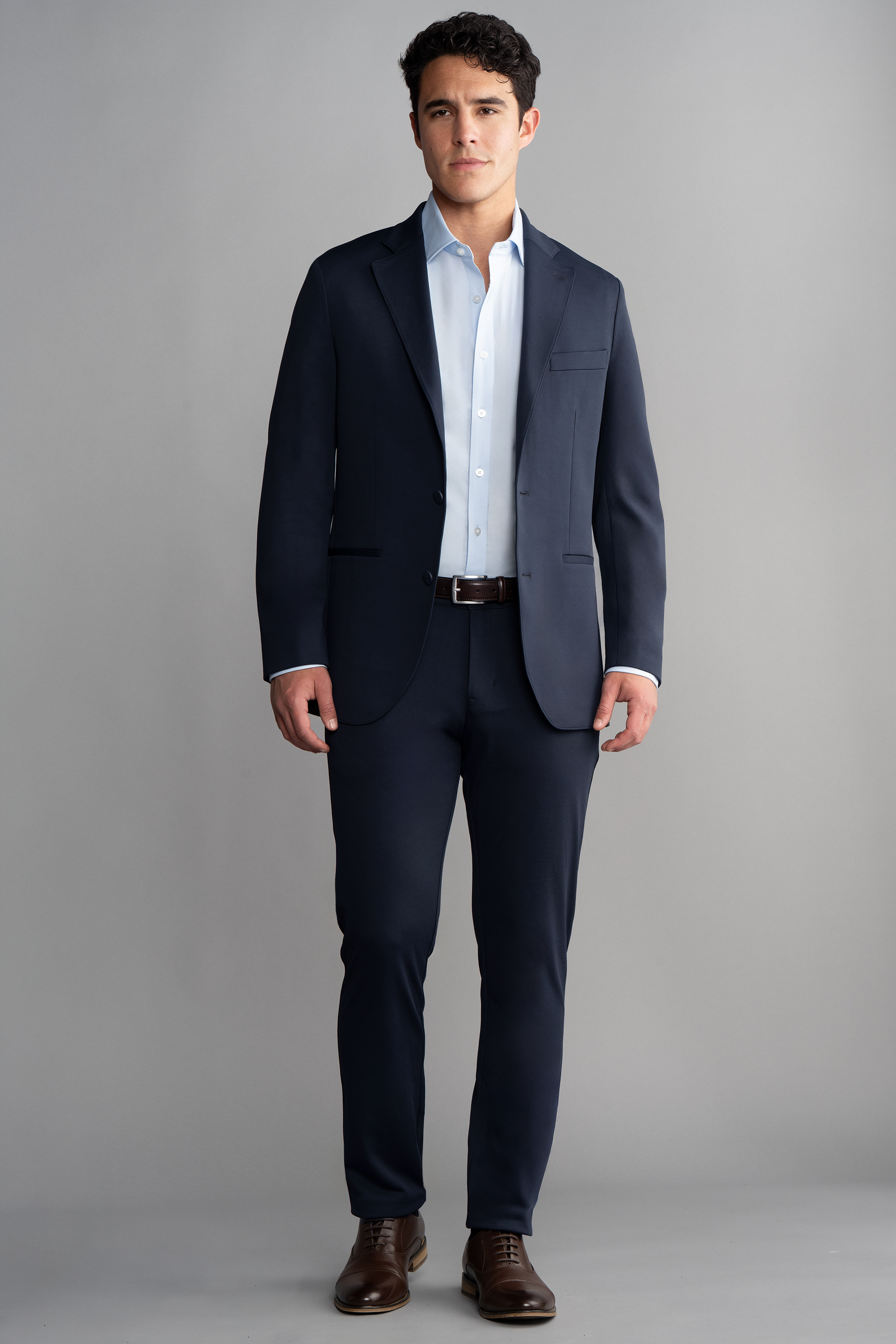 A man stands facing forward against a plain gray background, wearing the Twillory Circular Knit Blazer in Navy over a light blue dress shirt, paired with a brown belt and brown dress shoes. His expression is neutral with arms relaxed at his sides.