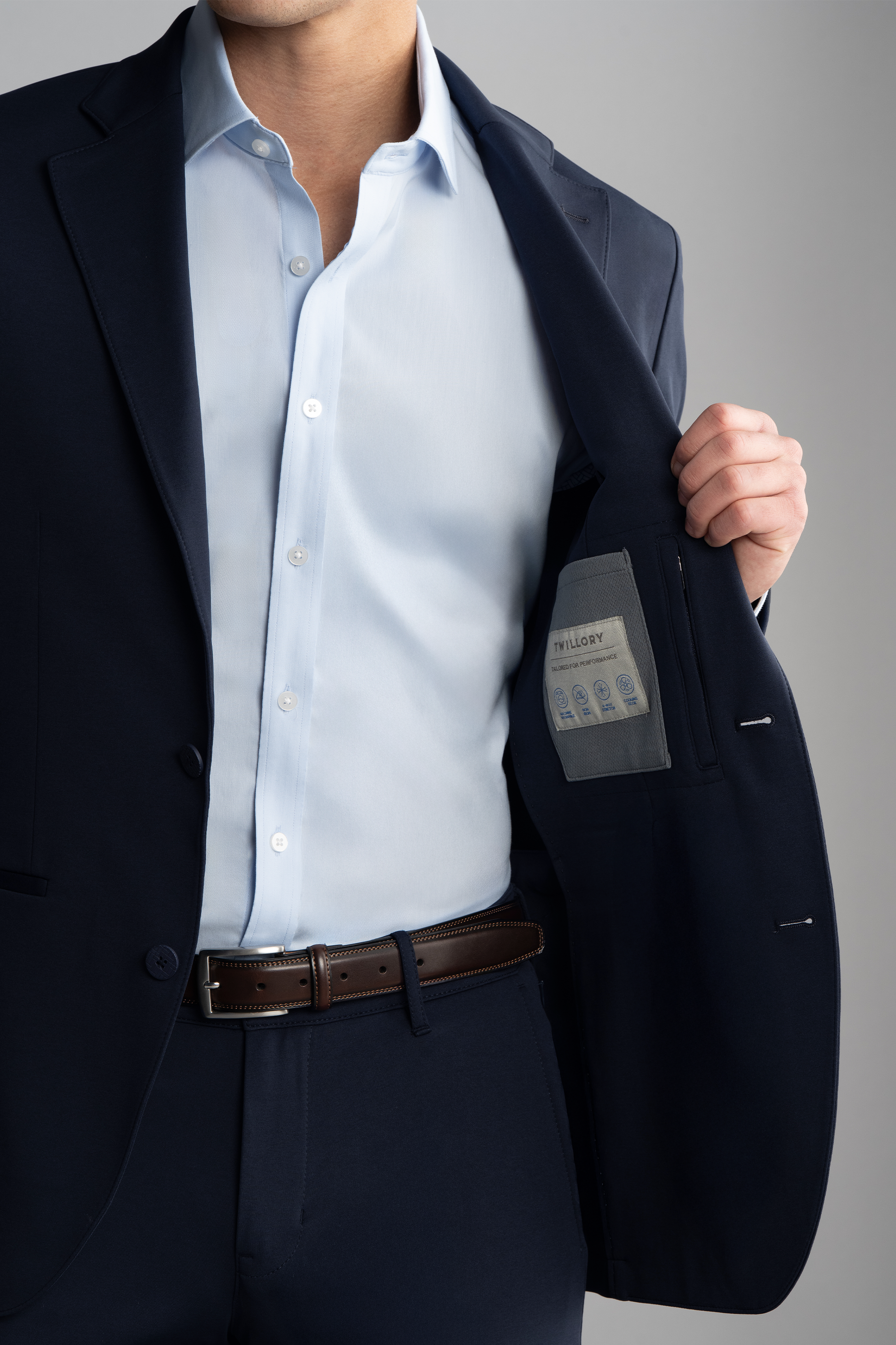 A man wearing the Twillory Circular Knit Blazer in Navy and a light blue shirt holds open his jacket to reveal the interior label. Only his torso is visible; he wears a brown leather belt and stands against a plain gray background.