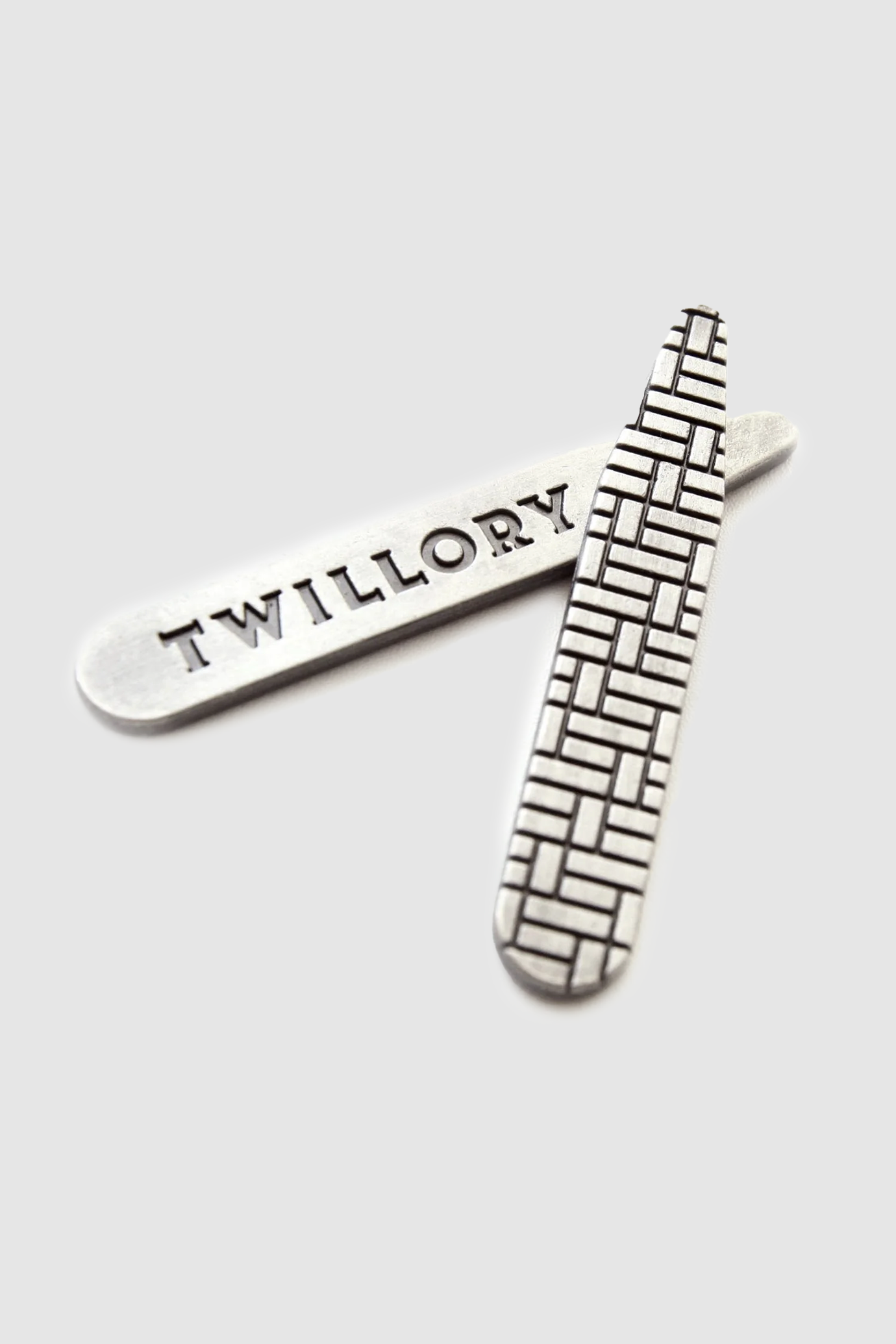 Two silver metal collar stays are displayed on a light gray backdrop beside the Twillory Leader Stripe // Micro Blue shirt. One stay is engraved with TWILLORY, the other features a geometric pattern. Both have rounded edges and a reflective finish.