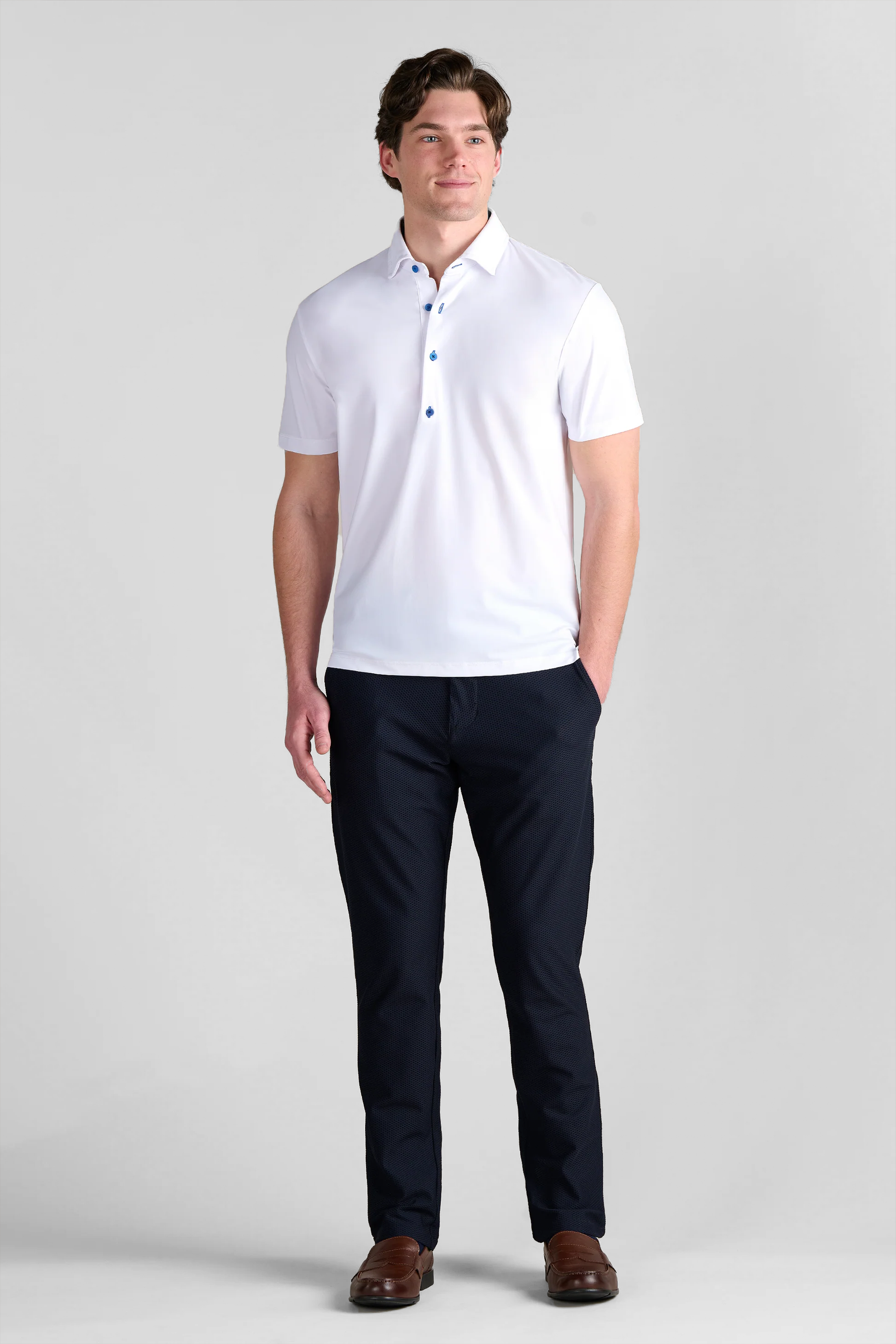 A man with short wavy brown hair smiles slightly, standing against a plain light gray background. He wears Twillory’s Event Polo Contrast in White Blue, navy pants, and brown loafers, with his hands in his pockets.