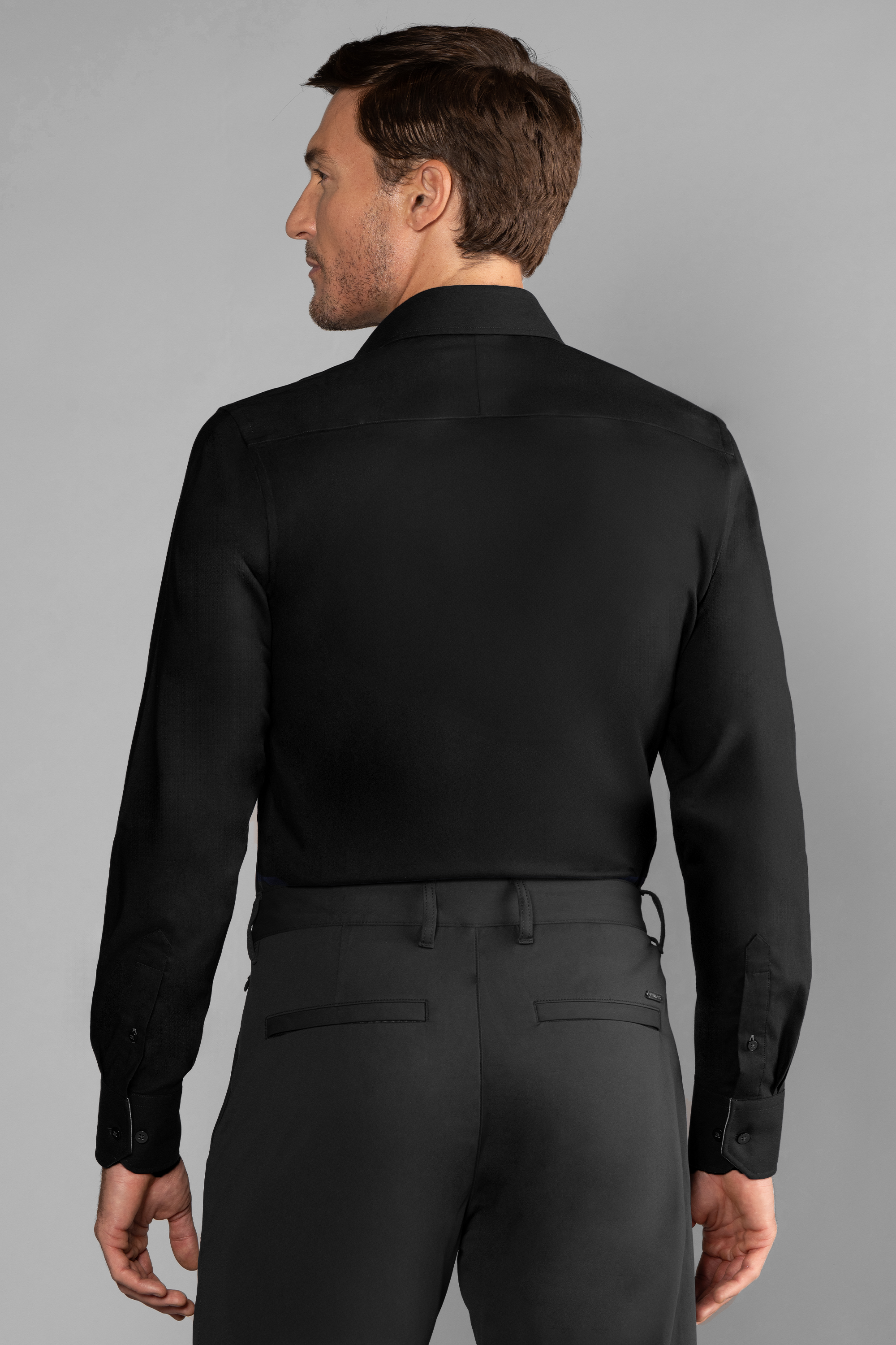 A man with short brown hair and a trimmed beard is seen from behind, wearing Twillory’s The Event Shirt // Black—an ideal wrinkle-free choice for men's officewear—paired with black dress pants against a plain light gray background.