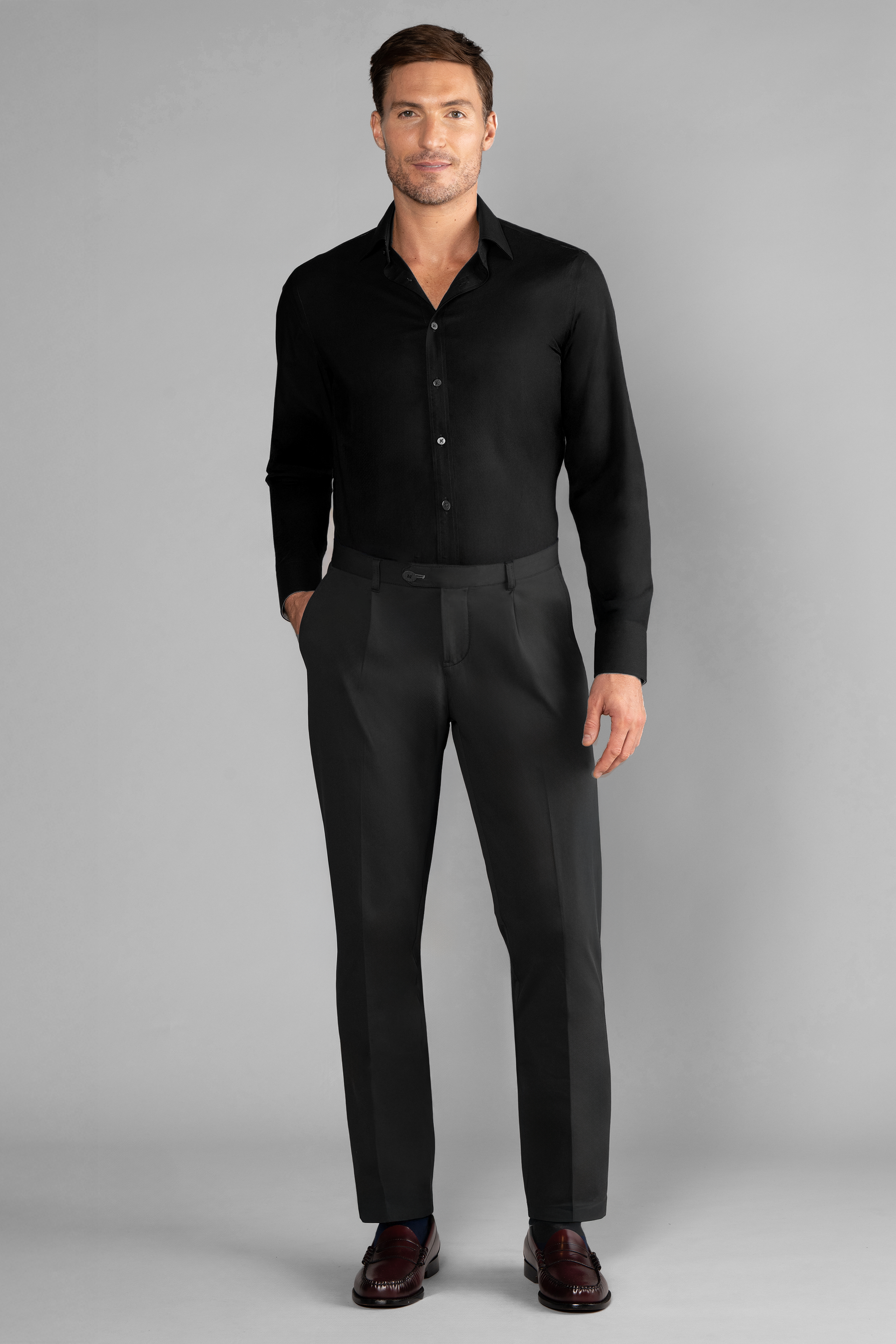 A man with short brown hair and a slight smile stands facing forward, one hand in his pocket, wearing Twillory's The Event Shirt // Black. He pairs it with dark dress pants and burgundy shoes against a plain light gray background—ideal officewear.