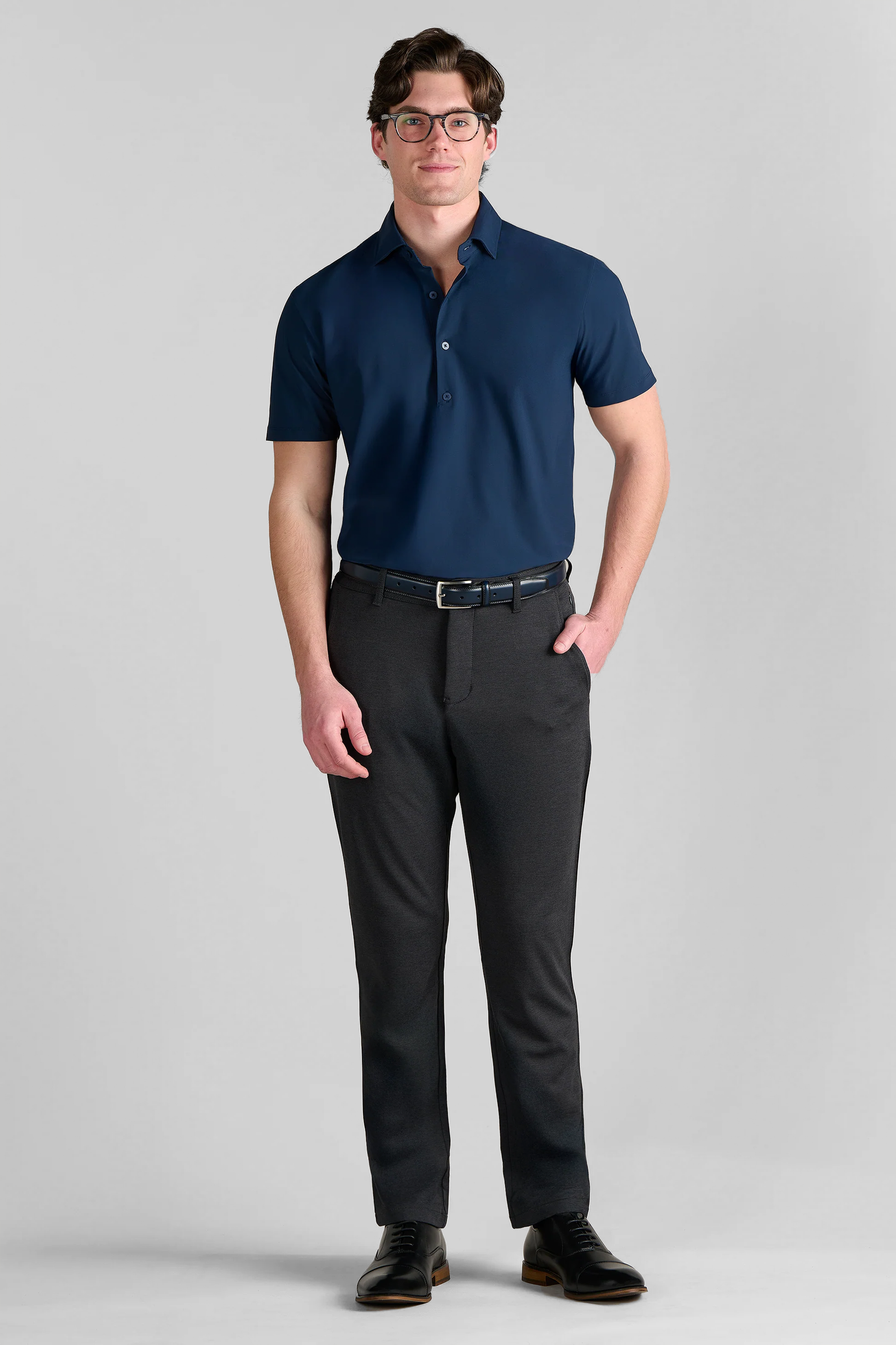 A man stands facing forward against a light gray background, wearing glasses and the Twillory Event Polo Solid in navy, paired with dark gray slacks, a black belt, and black dress shoes. One hand is in his pocket and he smiles slightly.