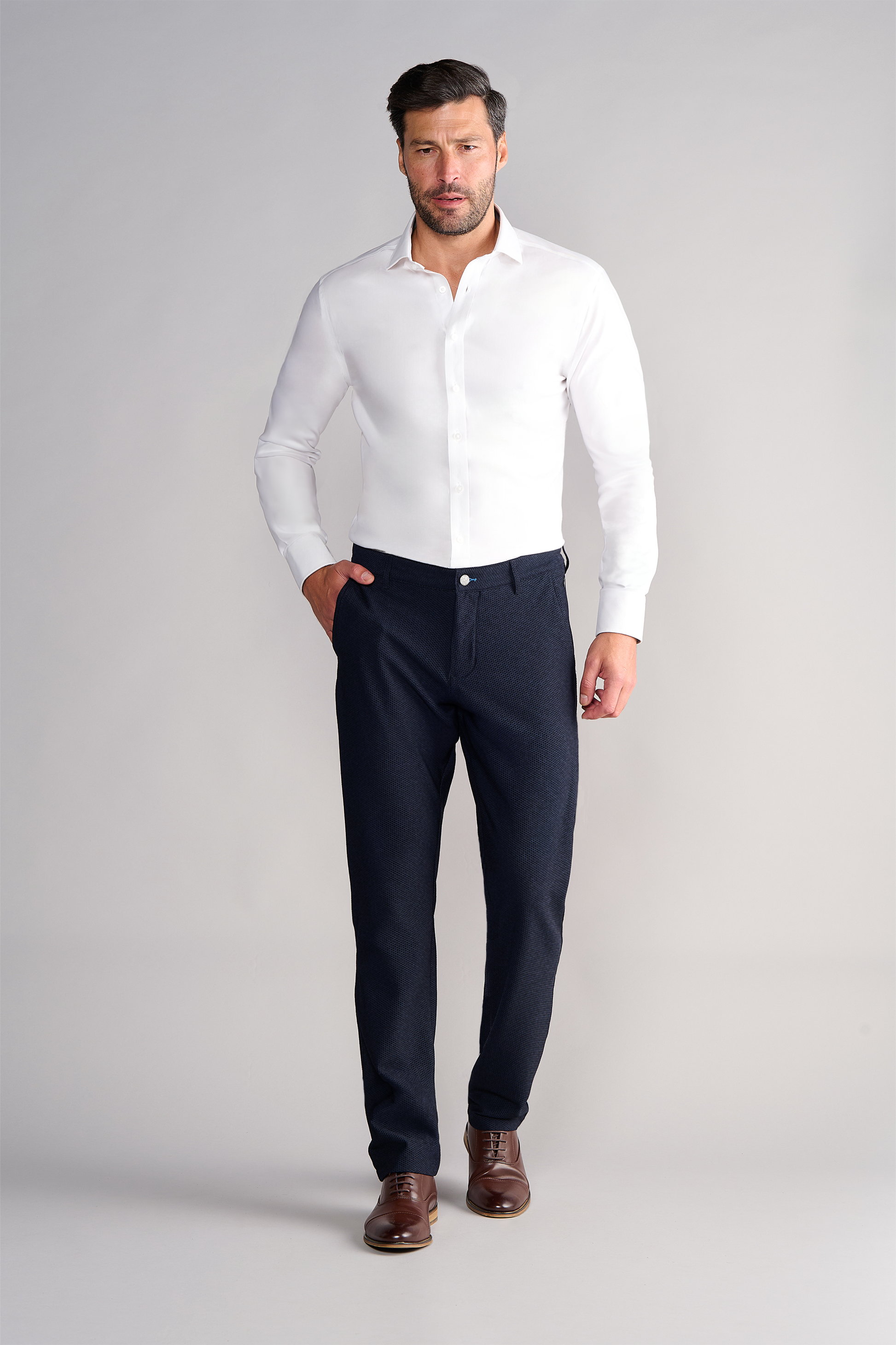 A man with short dark hair and a beard stands against a plain light background, wearing a fitted white shirt and Twillory AIR Pants in Heather Navy. He has one hand in his pocket and a neutral expression.