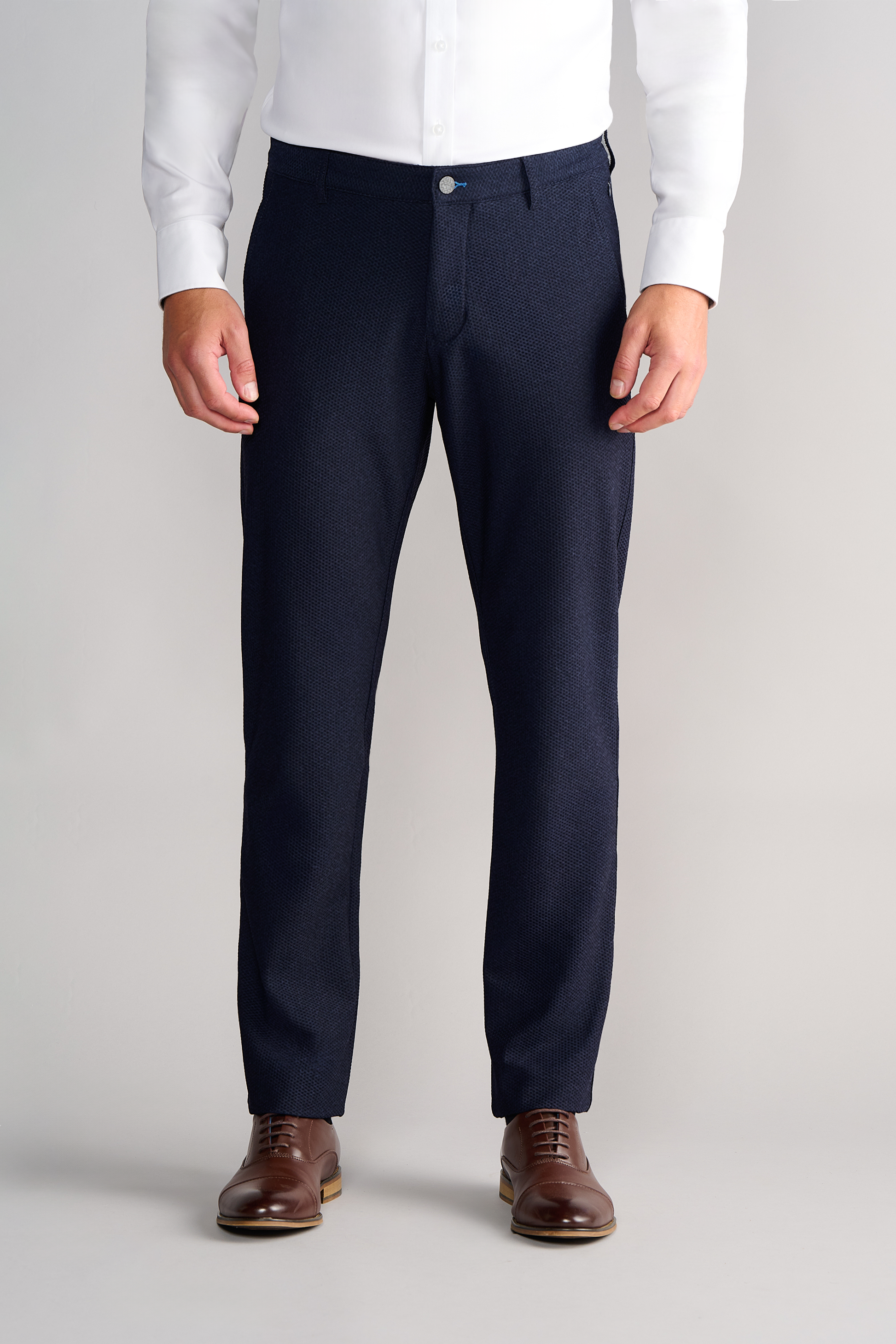 A man stands straight with arms relaxed, wearing a white dress shirt tucked into Twillory AIR Pants // Heather Navy and brown leather dress shoes. The pants are made from 360° Airflow Fabric. Background is neutral light gray.