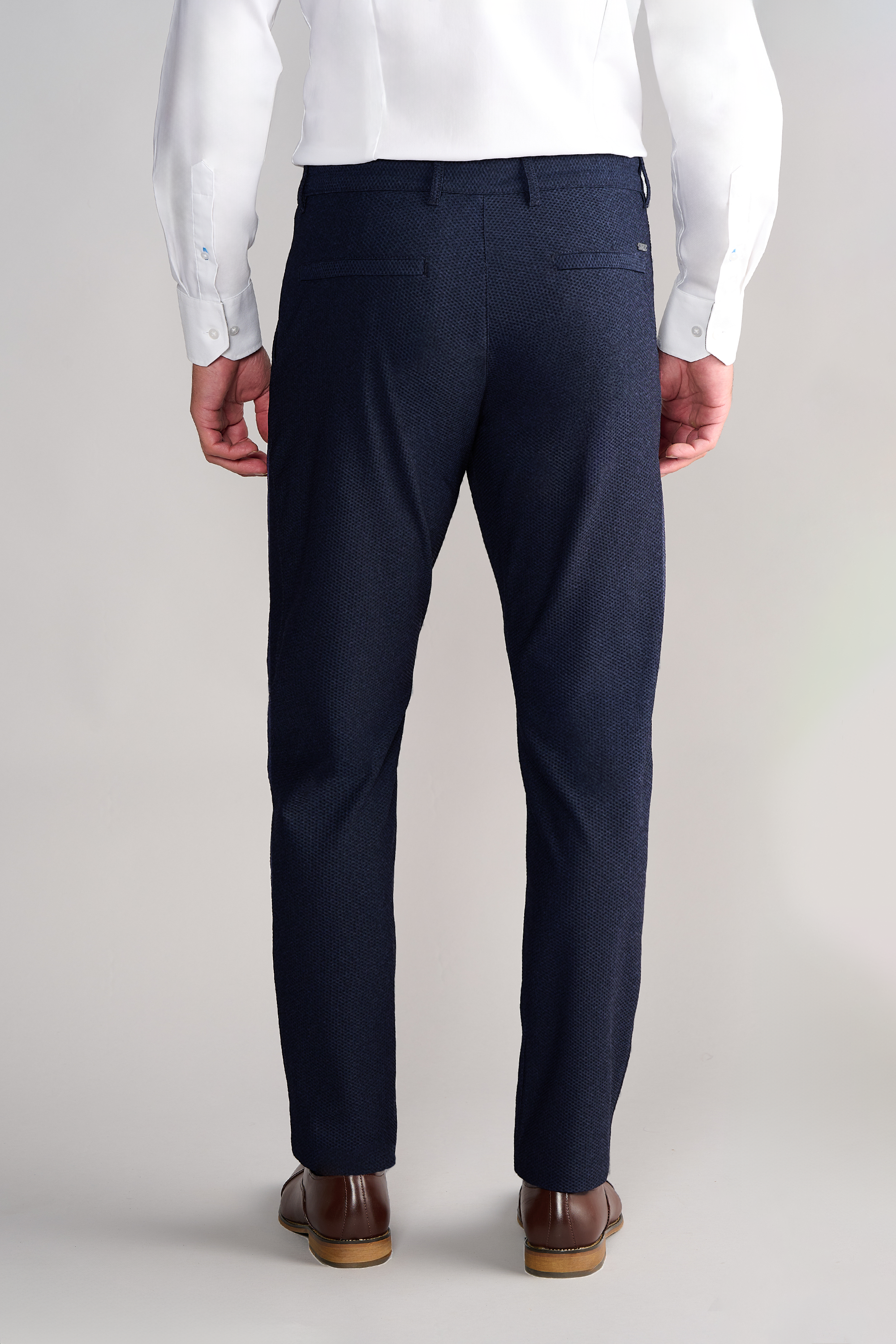 A man is seen from the back wearing Twillory AIR Pants // Heather Navy and a white dress shirt. The pants feature 360° Airflow Fabric, two back pockets with buttoned flaps. He stands on a light floor against a plain neutral background.