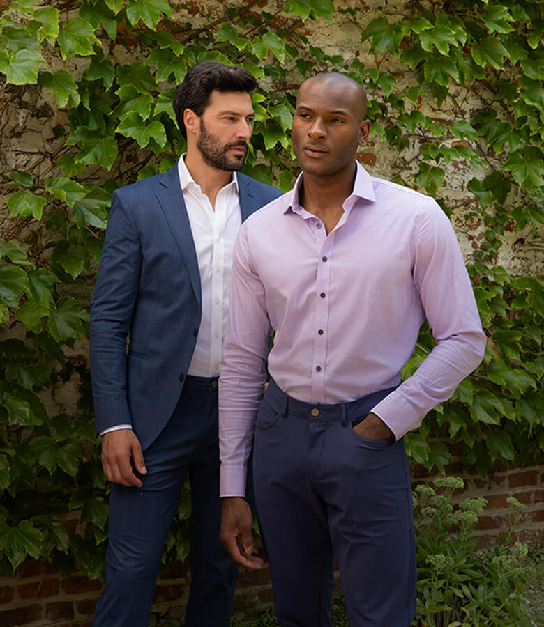 Two men wearing stylish formal attire, one in a blue suit and the other in a pink shirt, standing in front of a leafy garden backdrop.