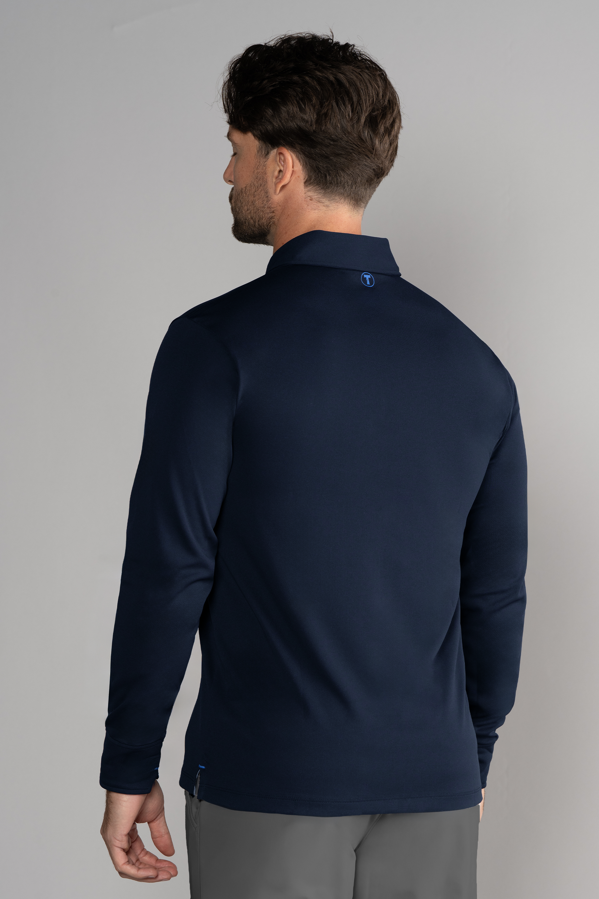 A man with short brown hair faces away from the camera, wearing the Twillory Long Sleeve Polo Solid in navy and light gray pants. The background is plain and light gray; his right arm hangs relaxed by his side.