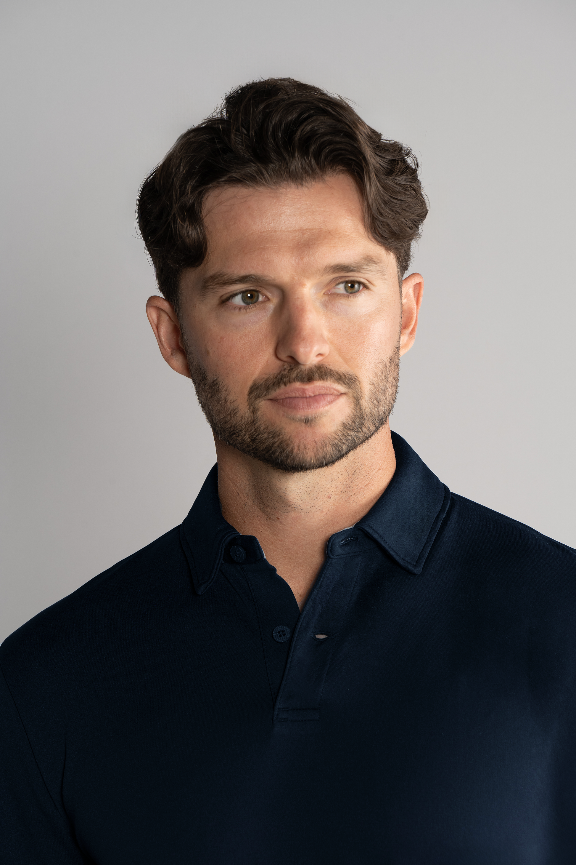A man with short, wavy brown hair and a trimmed beard wears the Twillory Long Sleeve Polo Solid in Navy. He faces slightly right, looking off-camera with a neutral expression against a plain light gray background.