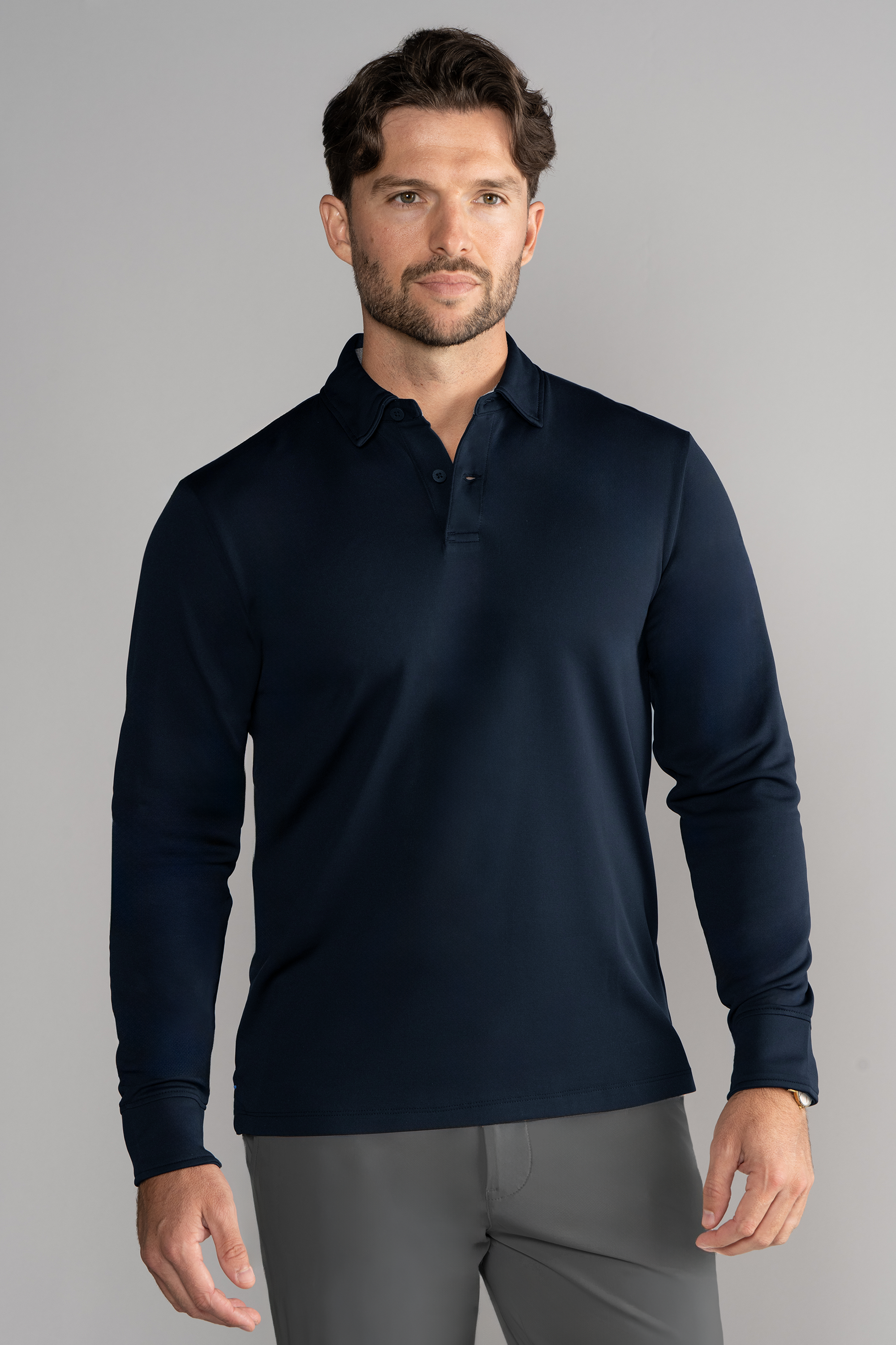 A man with short brown hair and light stubble wears a Twillory Long Sleeve Polo Solid in navy and gray pants. He stands facing forward against a plain light gray background with a neutral expression.