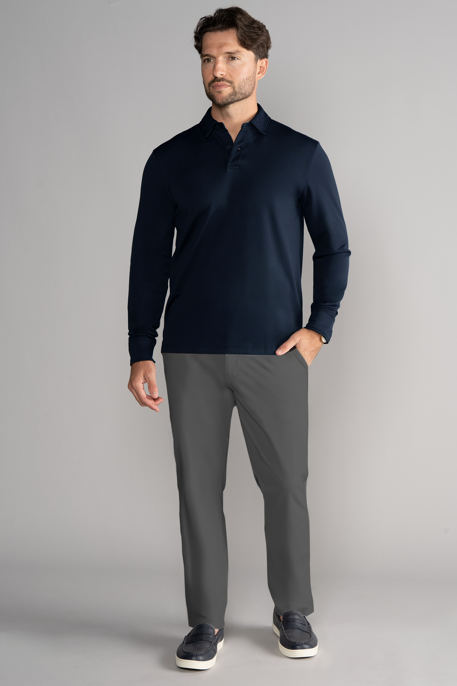 A man with short brown hair and a trimmed beard stands against a gray background, wearing the Twillory Long Sleeve Polo Solid in Navy, gray pants, and dark slip-on shoes with white soles, one hand in his pocket.