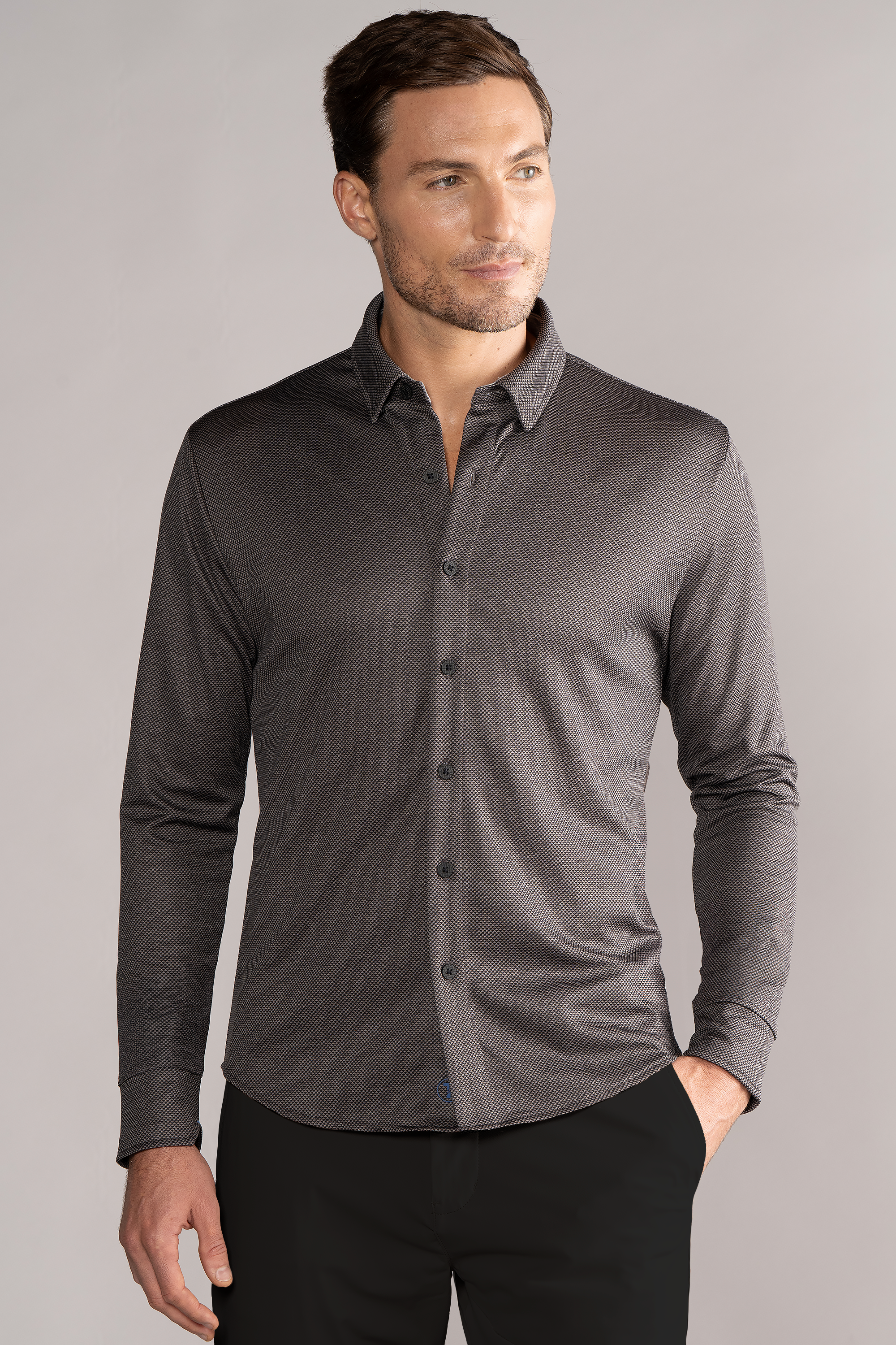 A light-skinned man with short brown hair wears a Twillory Long Sleeve Button Down Polo Dot in Grey Scale, untucked and slightly fitted. He stands with his left hand in his pocket against a plain light gray background.