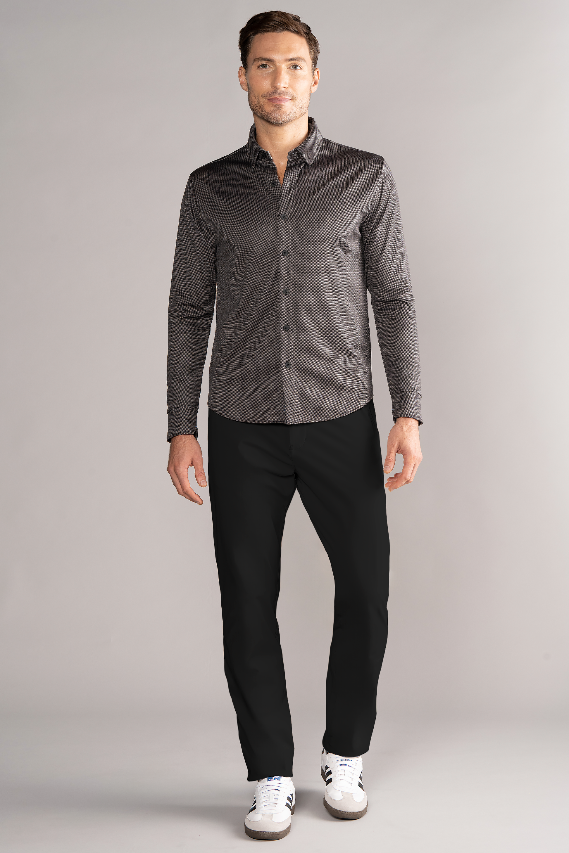 A man stands against a plain gray background, wearing the Twillory Long Sleeve Button Down Polo Dot in Grey Scale, paired with black pants and white sneakers. He has short brown hair, a trimmed beard, and a neutral expression.