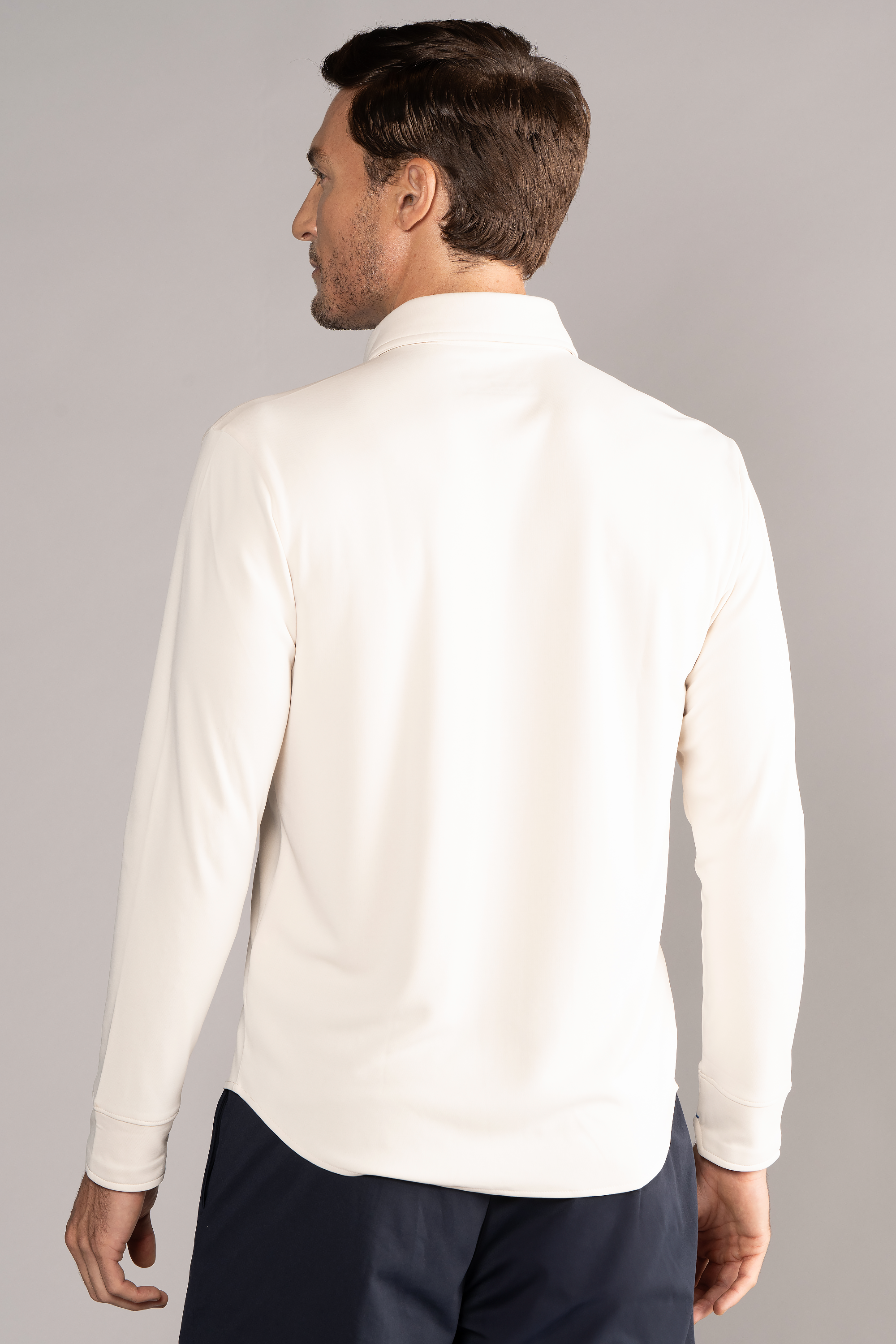 A man with short brown hair and a trimmed beard models the back of Twillory’s Long Sleeve Button Down Polo Solid in Ivory, paired with dark navy pants. The background is plain light gray with even lighting.