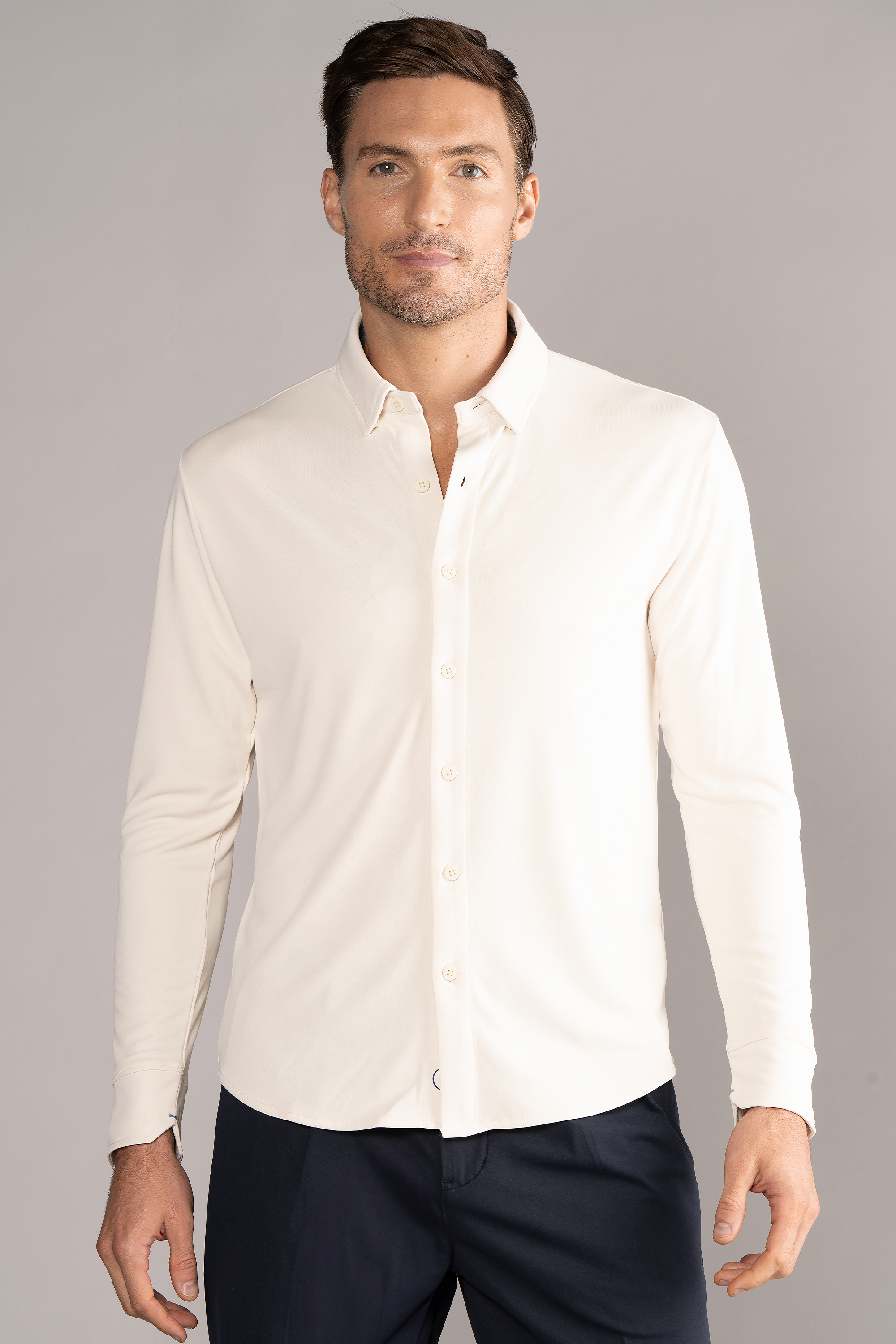 A man with short brown hair and light stubble wears the Twillory Long Sleeve Button Down Polo Solid in Ivory, paired with dark navy trousers. He stands against a plain light gray background, facing forward with a neutral expression and relaxed posture.