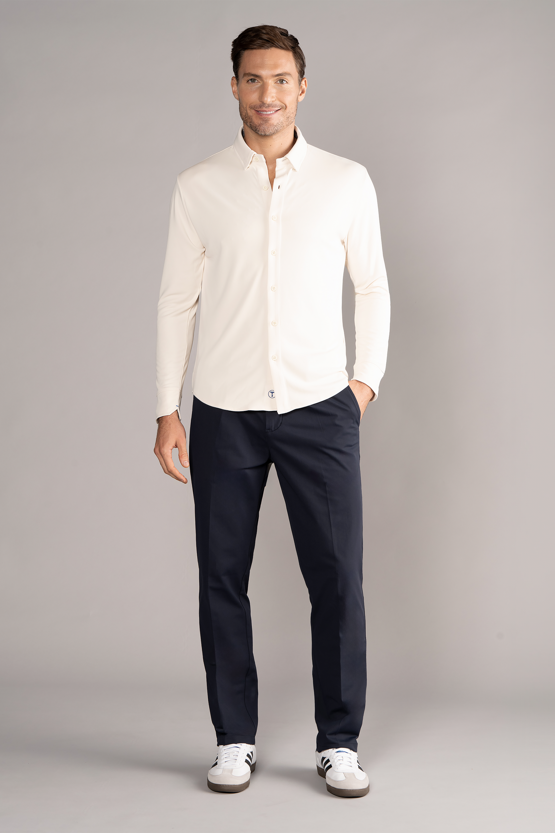A man stands against a plain gray background, smiling. He wears the Twillory Long Sleeve Button Down Polo Solid in Ivory with dark navy pants and white sneakers, hand in pocket for a casual yet polished look.