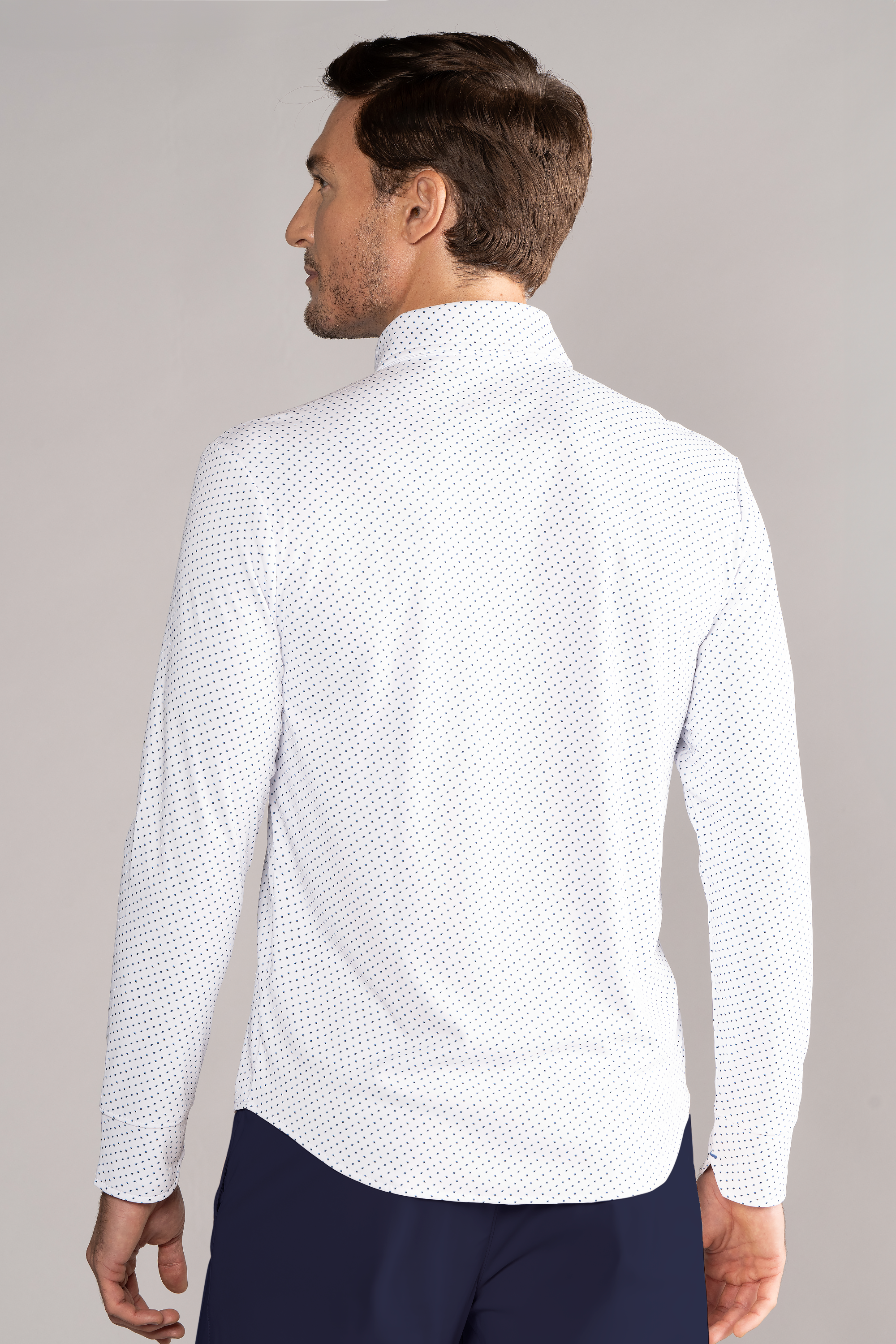 A man with short brown hair, seen from behind, wears the Twillory Long Sleeve Button Down Polo Dot // Blue Acorn shirt untucked over dark pants. The shirt features a small navy blue dot pattern. Background is plain light gray.