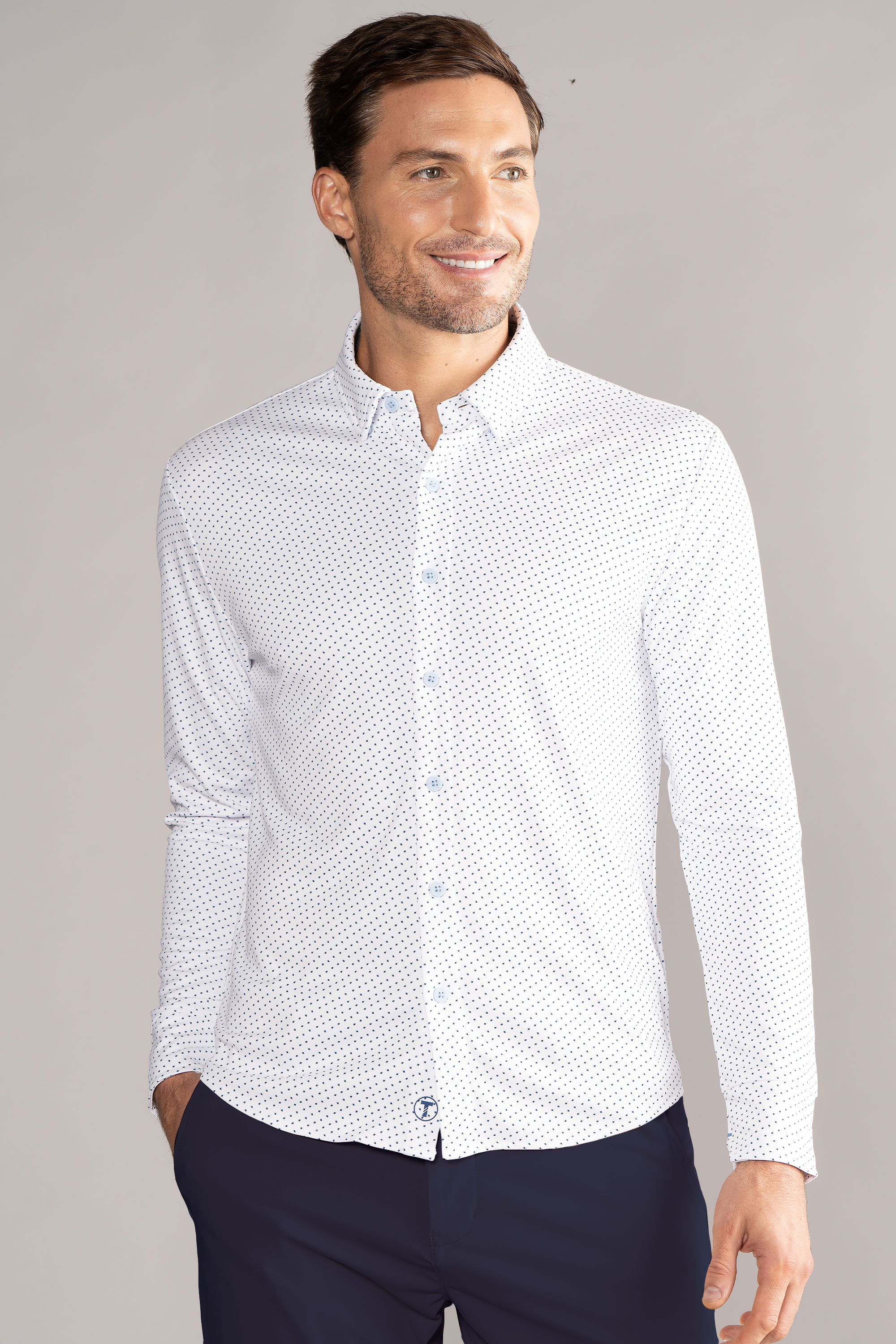 A man stands smiling with his left hand in his pocket, wearing Twillory's Long Sleeve Button Down Polo Dot // Blue Acorn and navy pants. He has neatly styled brown hair, and the background is plain light gray.