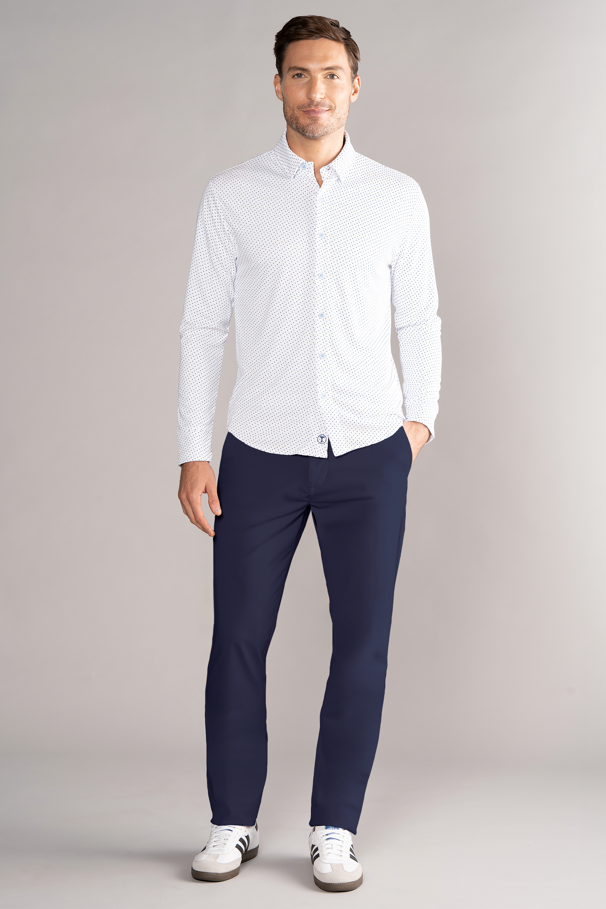 A man stands before a gray background, wearing Twillory’s Long Sleeve Button Down Polo Dot // Blue Acorn, paired with dark navy trousers and white sneakers with black stripes. He has short brown hair, a light beard, one hand in his pocket, and smiles slightly.