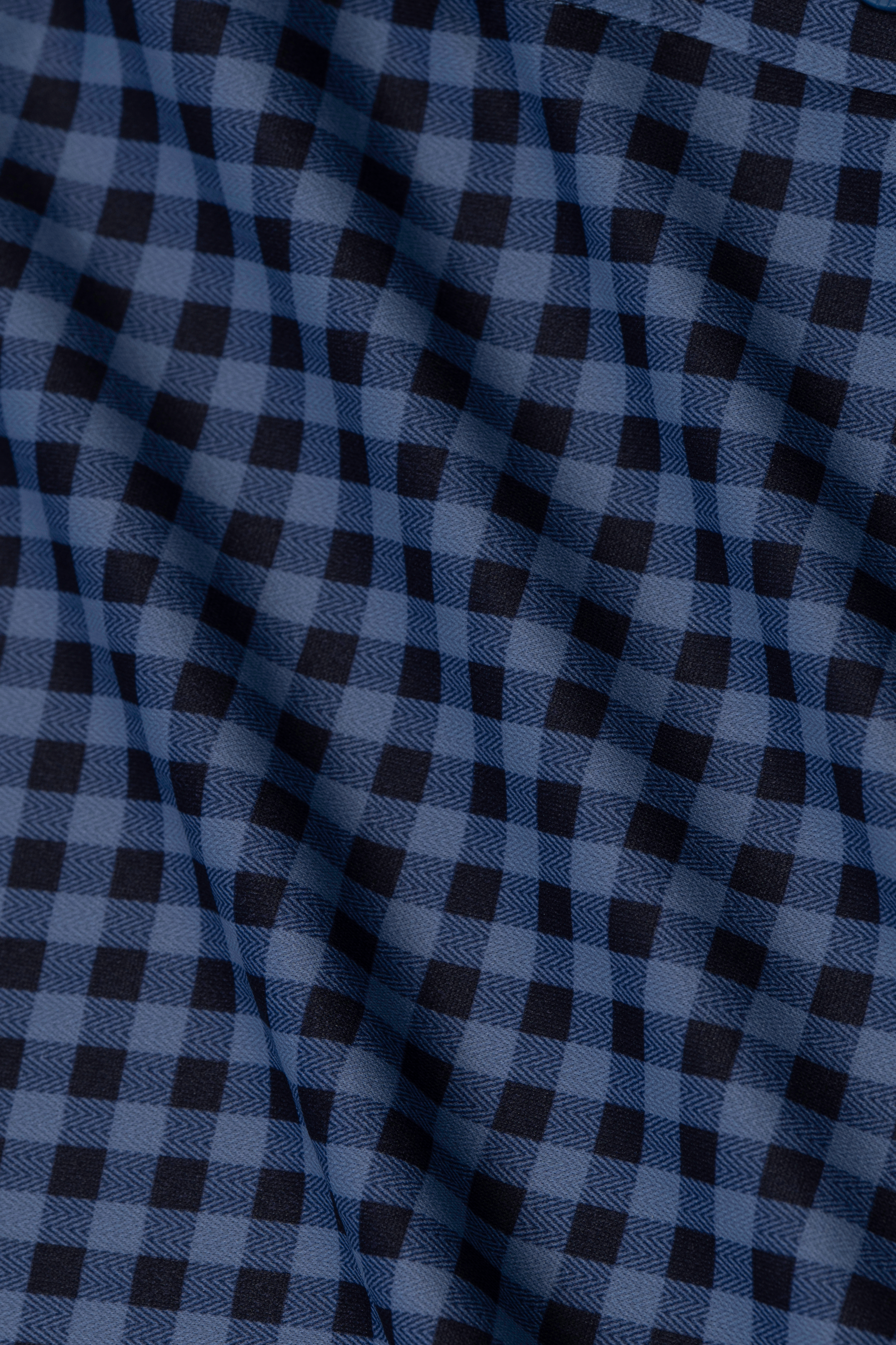 A close-up of wrinkle-free fabric from the Twillory Long Sleeve Button Down Polo Gingham in Navy Blue shows black and navy checks in a regular grid, with gentle folds and shadows highlighting its classic, textured look.