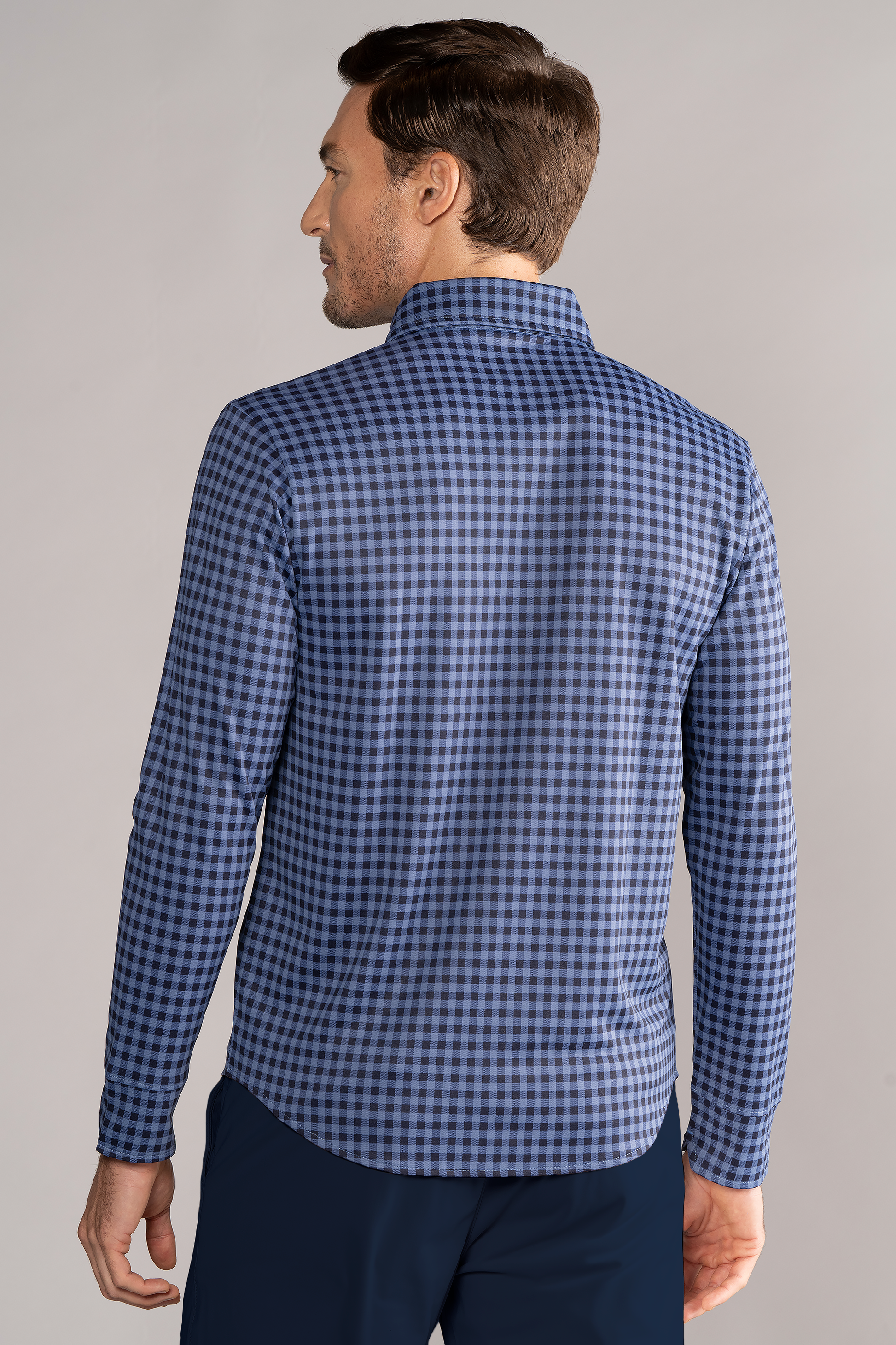 A man with short brown hair is seen from behind wearing the Twillory Long Sleeve Button Down Polo Gingham in Navy Blue, paired with dark blue pants, standing against a plain light gray background, his left arm relaxed at his side.