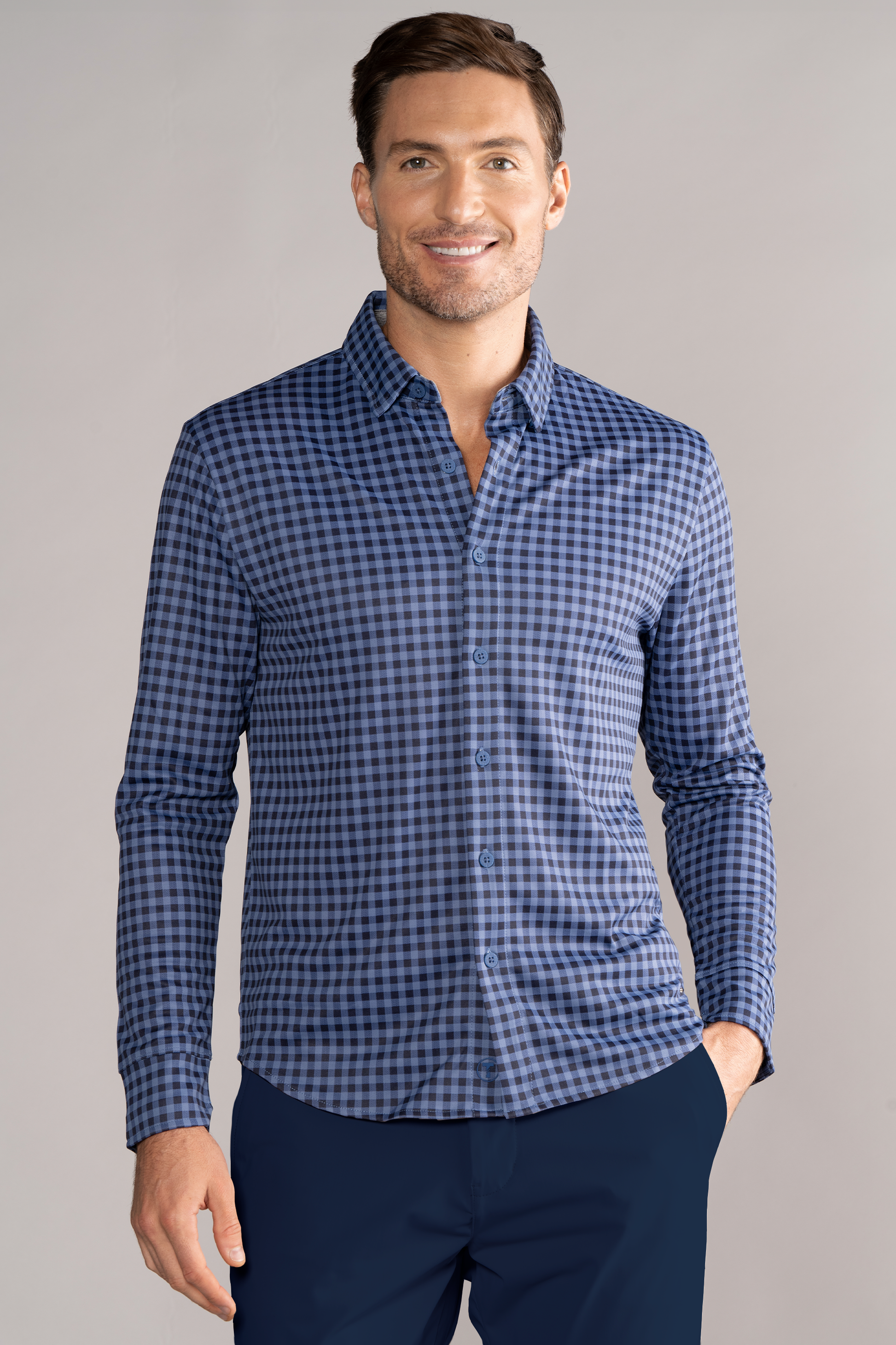 A man smiles with one hand in his pocket, wearing Twillory's Long Sleeve Button Down Polo Gingham in Navy Blue and dark blue pants, set against a plain light grey background.