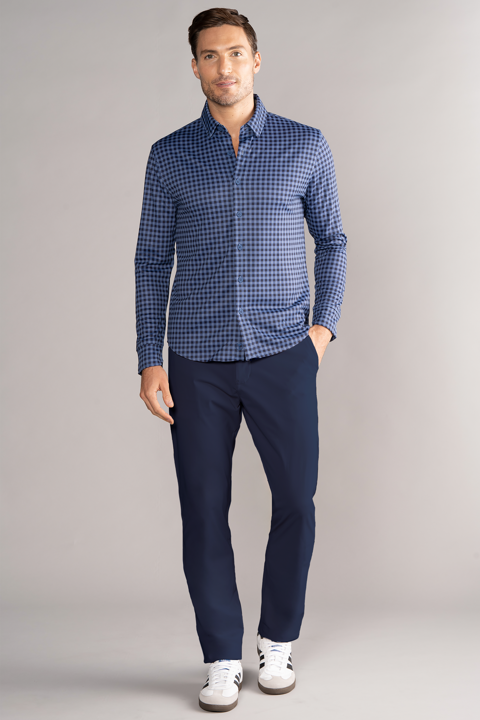 A man stands against a light gray background wearing the Twillory Long Sleeve Button Down Polo Gingham in Navy Blue, paired with dark blue pants and white sneakers. He has short brown hair, a slight beard, and one hand in his pocket.
