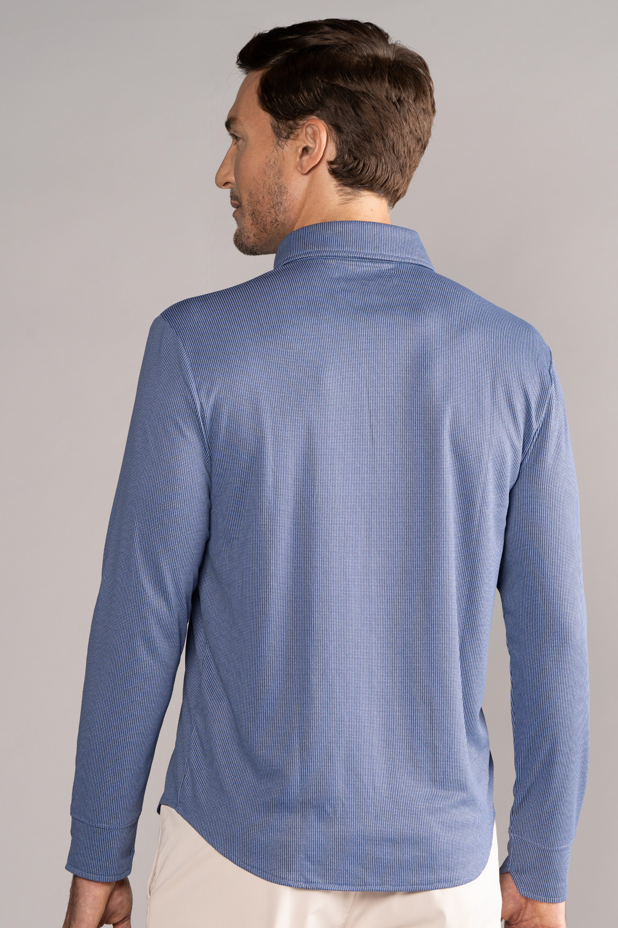 A man with short brown hair is seen from behind, wearing the Twillory Long Sleeve Button Down Polo Dot in Blue Puppytooth. He stands with relaxed arms against a plain, light grey background.