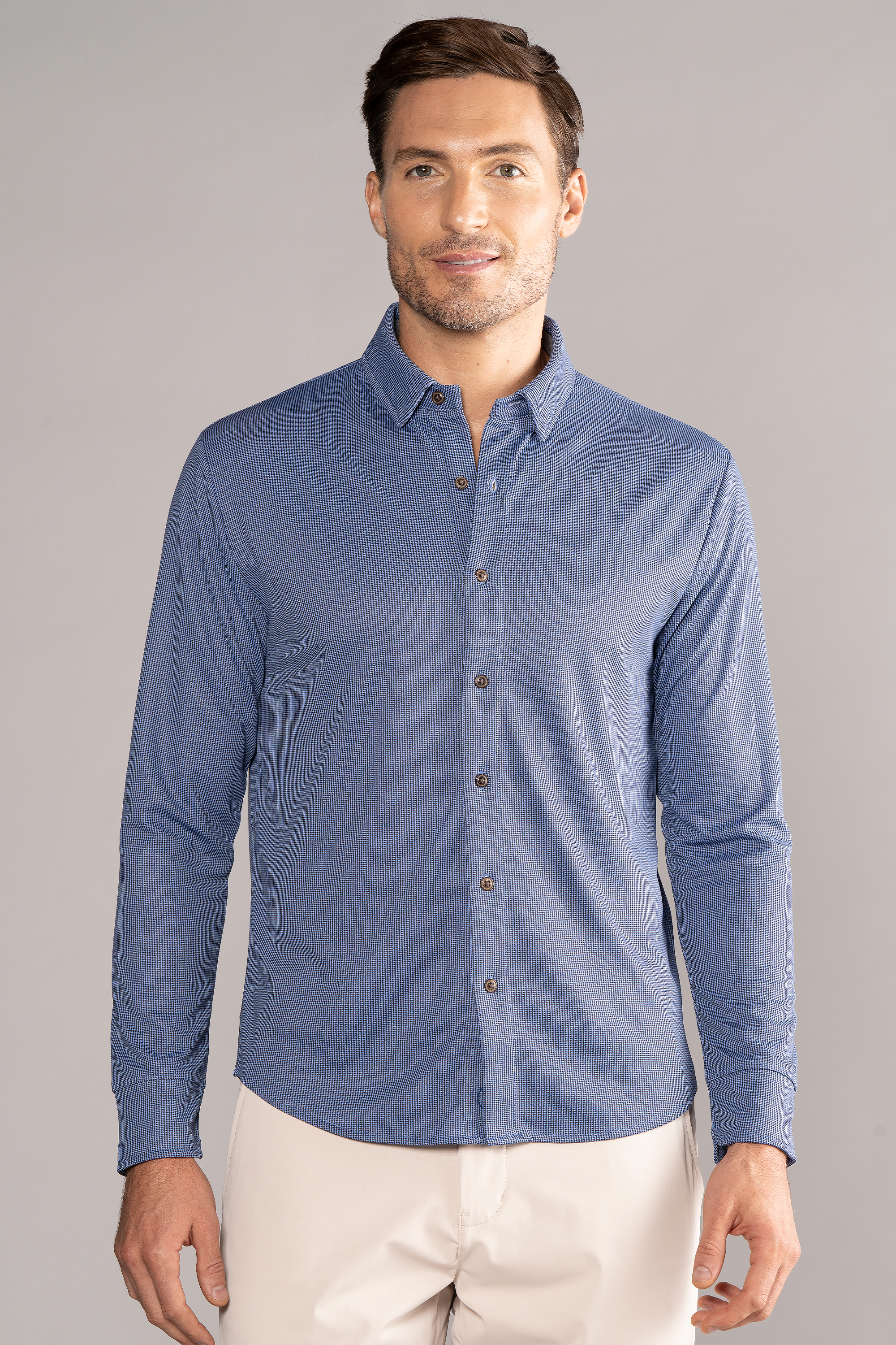 A man with short brown hair and light facial hair stands facing forward against a plain, light grey background, smiling slightly while wearing the Twillory Long Sleeve Button Down Polo Dot in Blue Puppytooth.