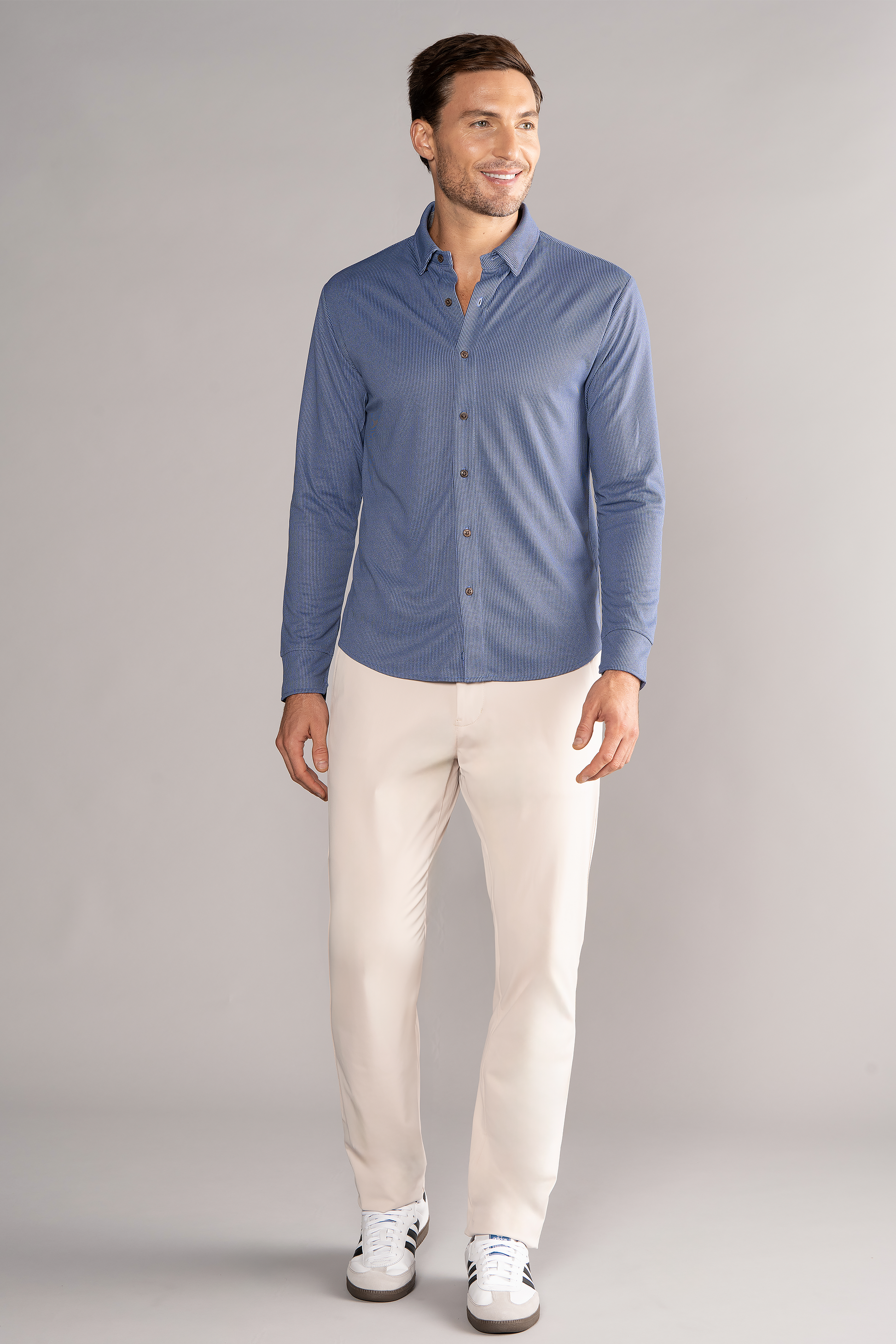 A man with short brown hair and a neat beard stands smiling in a Twillory Long Sleeve Button Down Polo Dot shirt in Blue Puppytooth, paired with light beige pants and white sneakers with black accents against a gray background.