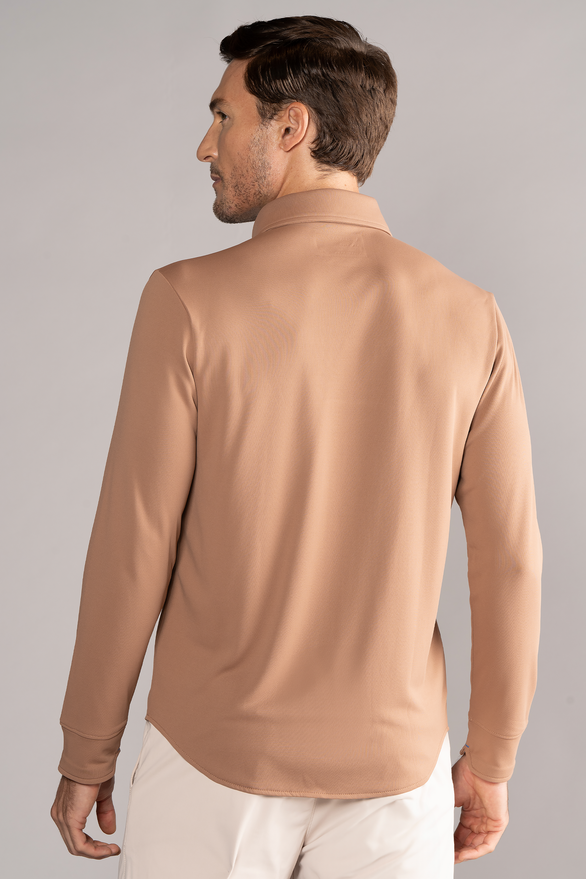 A man with short brown hair is seen from the back, wearing the Twillory Long Sleeve Button Down Polo Solid in Clay with a relaxed fit, paired with white pants. He stands against a plain light gray background, facing slightly left.