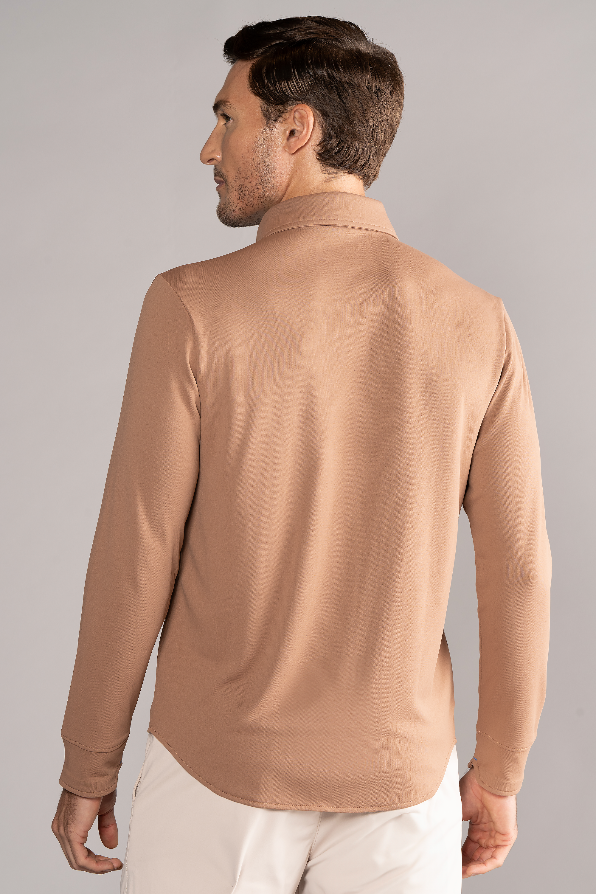 A man with short brown hair is seen from the back, wearing the Twillory Long Sleeve Button Down Polo Solid in Clay with a relaxed fit, paired with white pants. He stands against a plain light gray background, facing slightly left.