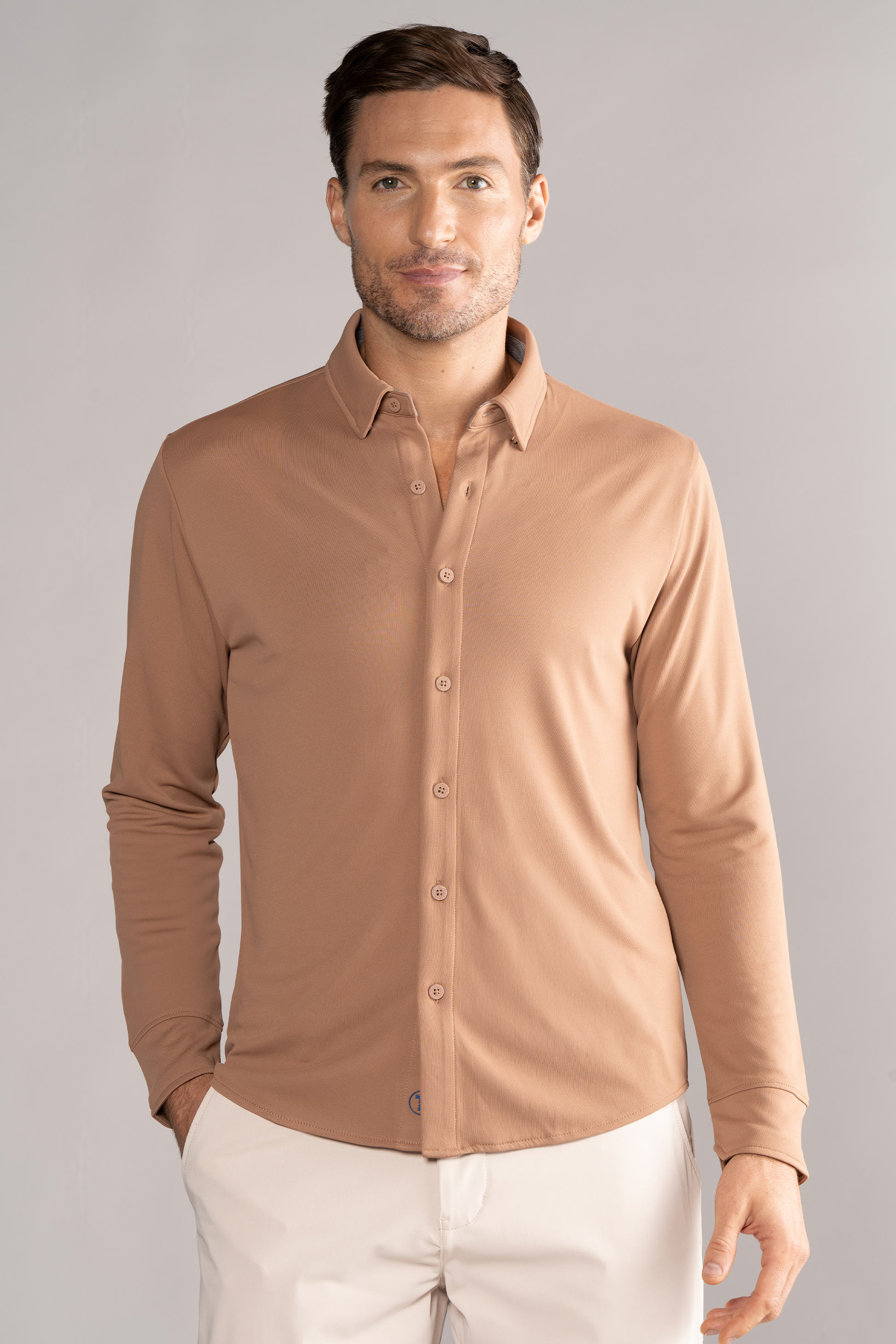 A man with short brown hair and light skin wears the Twillory Long Sleeve Button Down Polo Solid in Clay and beige pants. He stands facing forward, smiling gently with his hands in pockets, against a plain light grey background.