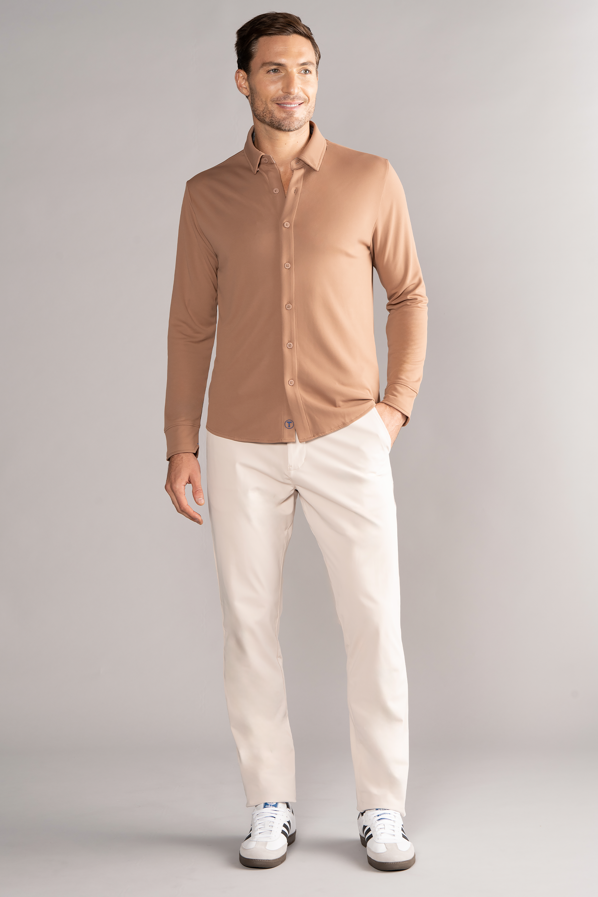 A man stands smiling in front of a gray background, wearing the Twillory Long Sleeve Button Down Polo Solid in Clay. His right hand rests by his side, and his left hand is partially in his pocket.
