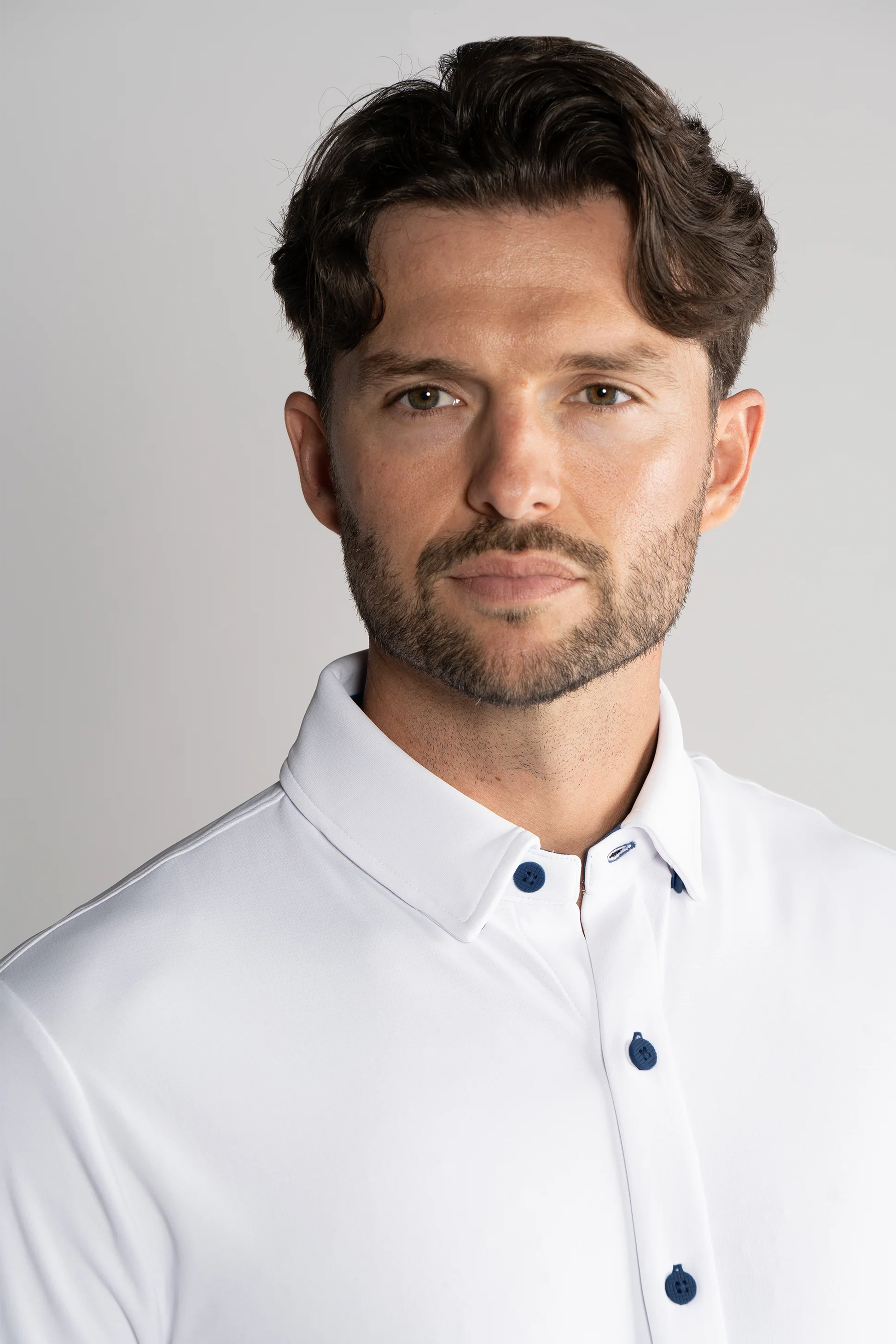 A man with wavy brown hair and a short beard wears the Twillory Long Sleeve Button Down Polo Contrast in White Navy, looking at the camera with a neutral expression against a light gray background. The photo is from the chest up.